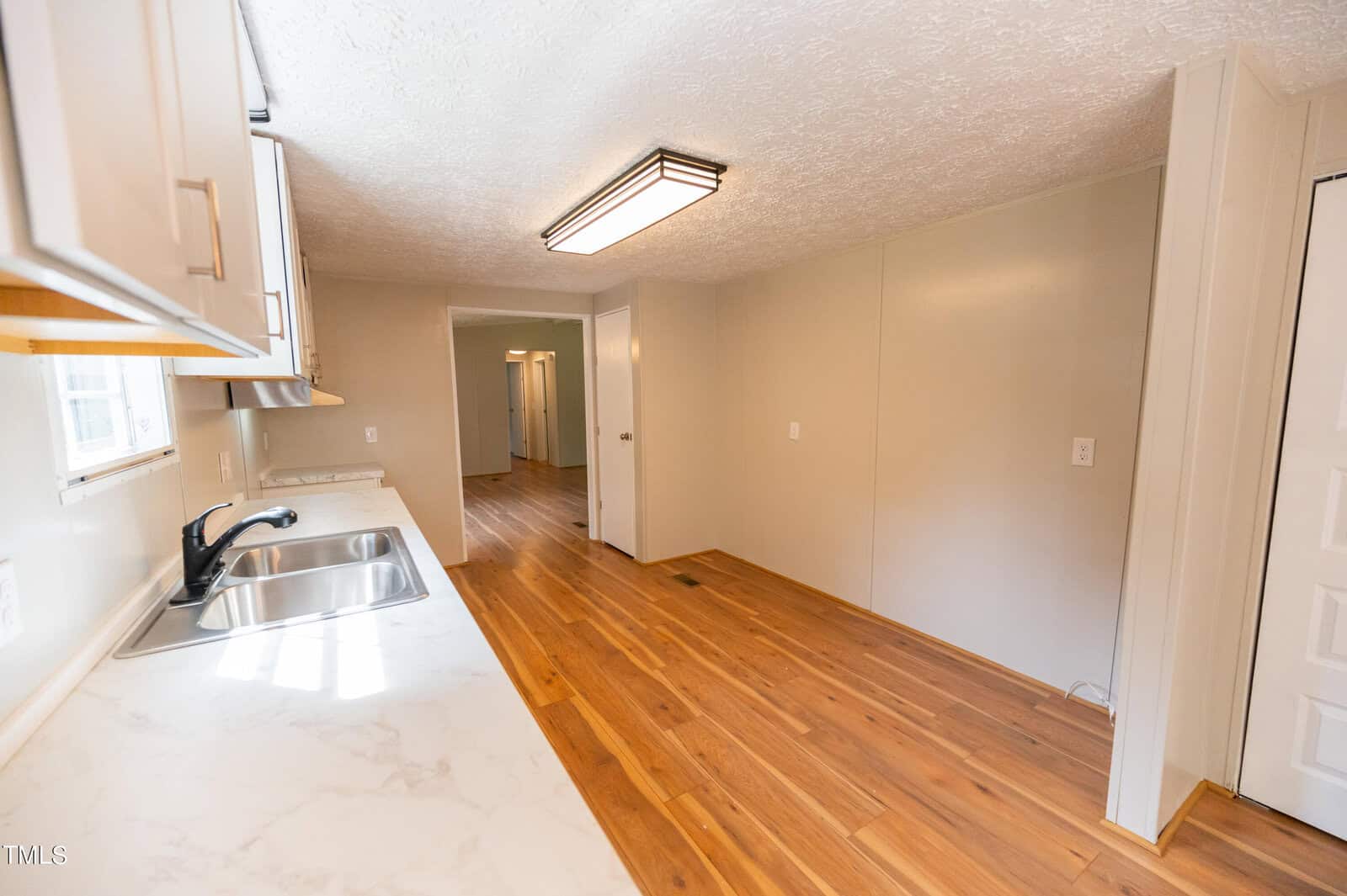 A narrow kitchen with white countertops and light brown wooden cabinets. A black sink and modern faucet are visible on the left. The room features light-colored walls, a wood-style floor, and recessed ceiling lighting. A hallway leads to additional rooms.