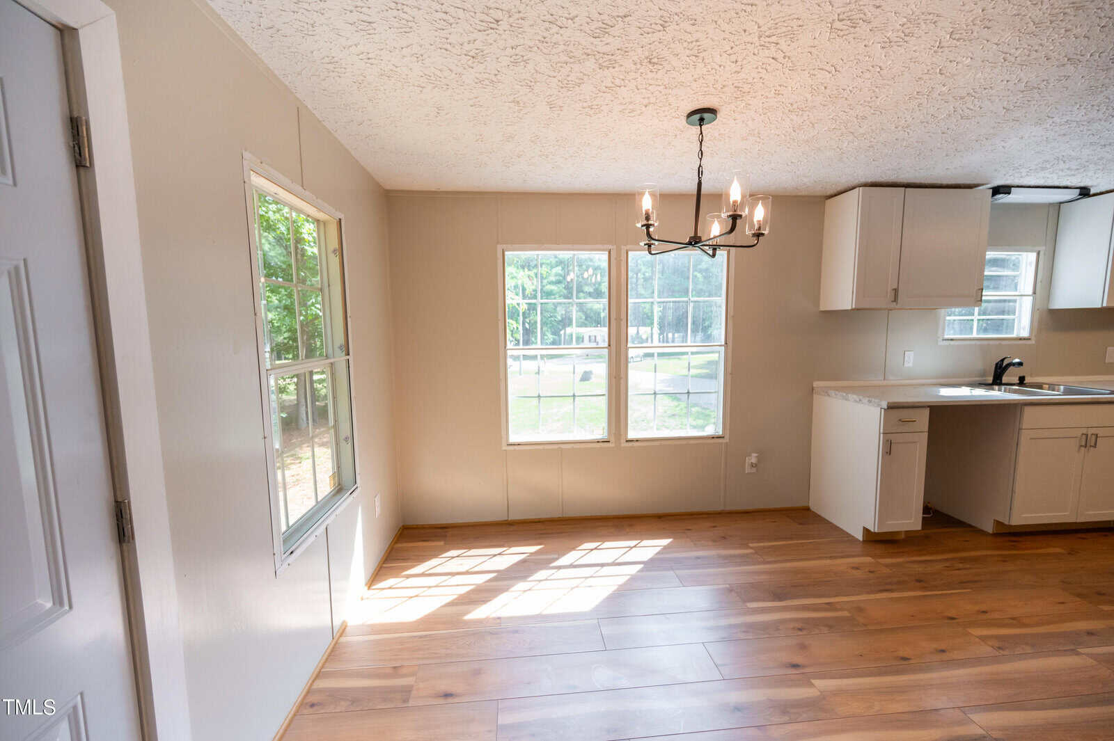 A bright, empty room with large windows letting in natural light. The room has light-colored walls, a wood-patterned floor, and a single light fixture hanging from the ceiling. The space appears to be a combination of dining and kitchen areas.