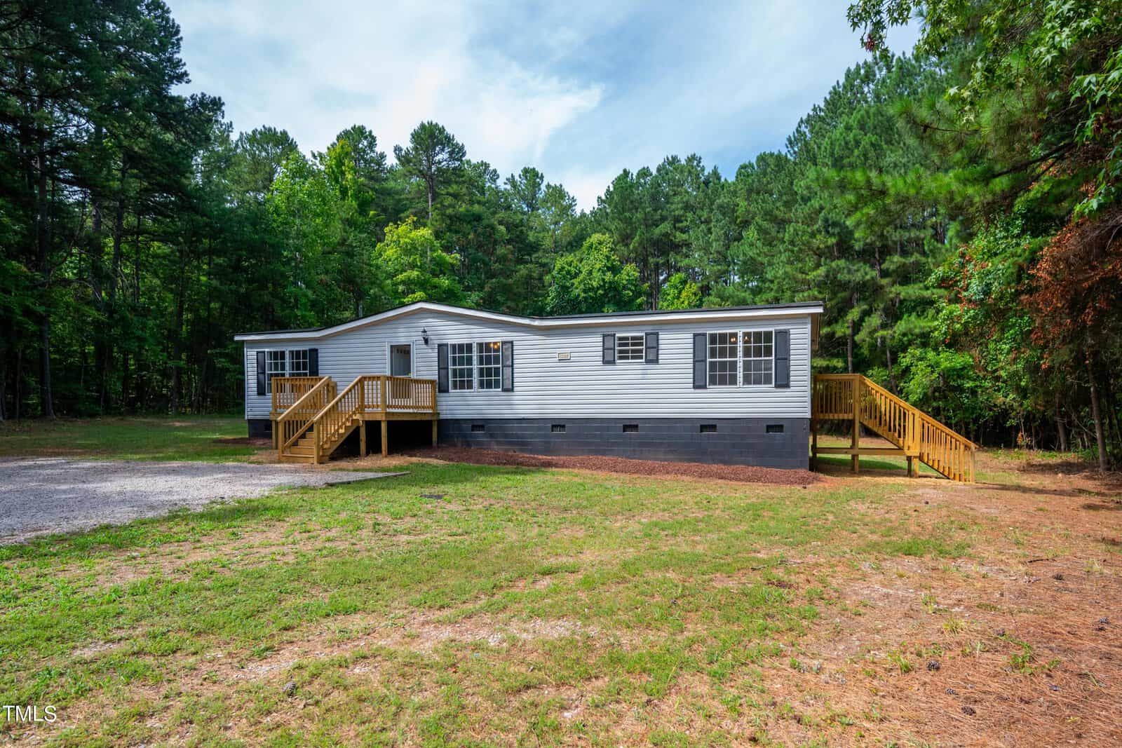 A single-story manufactured home at 42 Canary Lane, Henderson NC, with light gray siding and white trim features an extended front porch with steps and wooden railings. The house is set in a clearing surrounded by tall, dense trees, with a grassy yard and a gravel driveway.