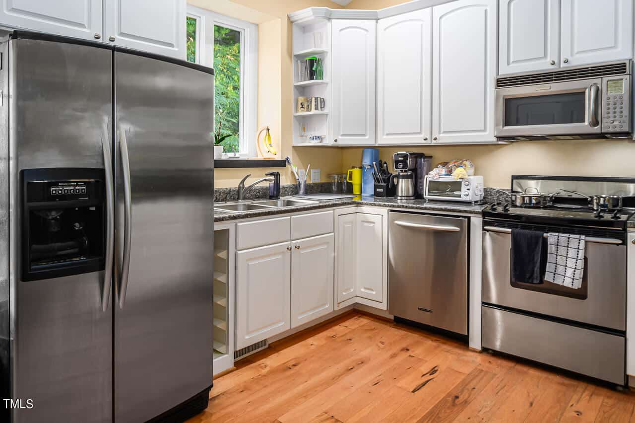 A modern kitchen with white cabinets, stainless steel appliances including a fridge, dishwasher, oven, and microwave. A window above the sink lets in natural light. Wooden floor and granite countertops add warmth to the space.