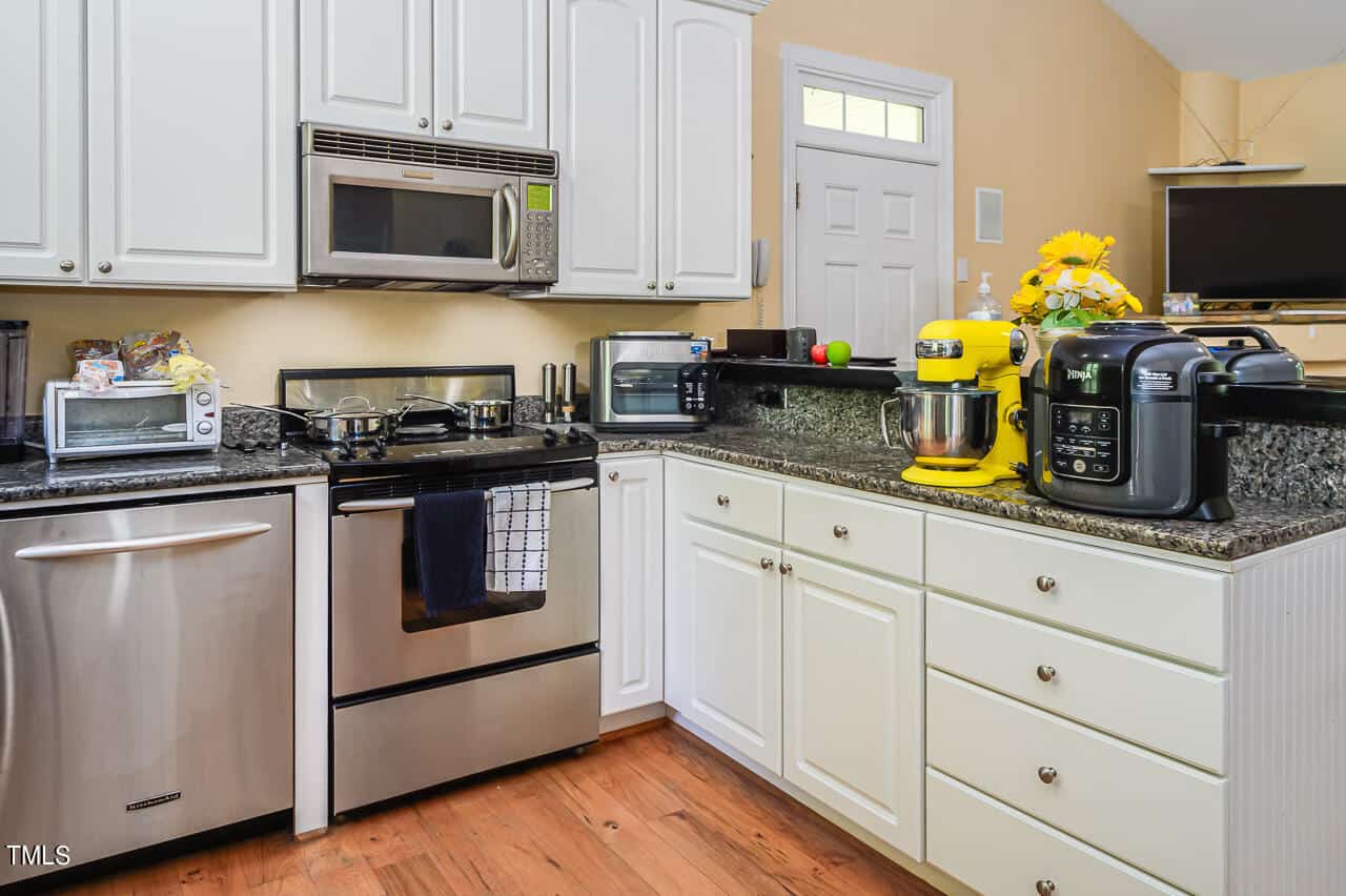 A modern kitchen with stainless steel appliances, including an oven, microwave, and dishwasher. Countertops are granite with small appliances like a toaster, mixer, blender, and air fryer. Cabinets are white, and there's a yellow wall in the background.