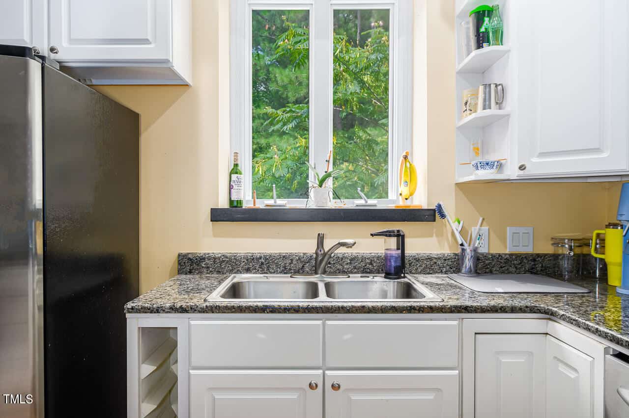 A kitchen scene featuring white cabinets, a stainless steel double sink, a black granite countertop, and a window showing green foliage outside. Various kitchen items are neatly placed, including a dish soap bottle and a banana on a stand.