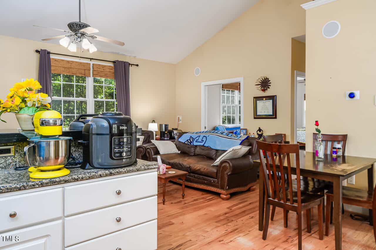 A cozy kitchen and living area featuring a granite countertop with a stand mixer and pressure cooker. A dining table with a flower vase sits nearby, while a sofa is visible in the background under large windows with natural light.