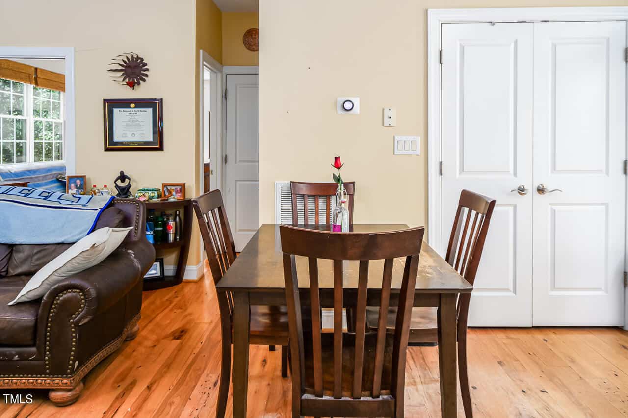 A cozy dining area with a wooden table and four chairs. A single red rose in a vase is on the table. The room has wooden flooring, a brown leather sofa, a wall with a diploma, and a door in the background. Natural light filters in from a window.