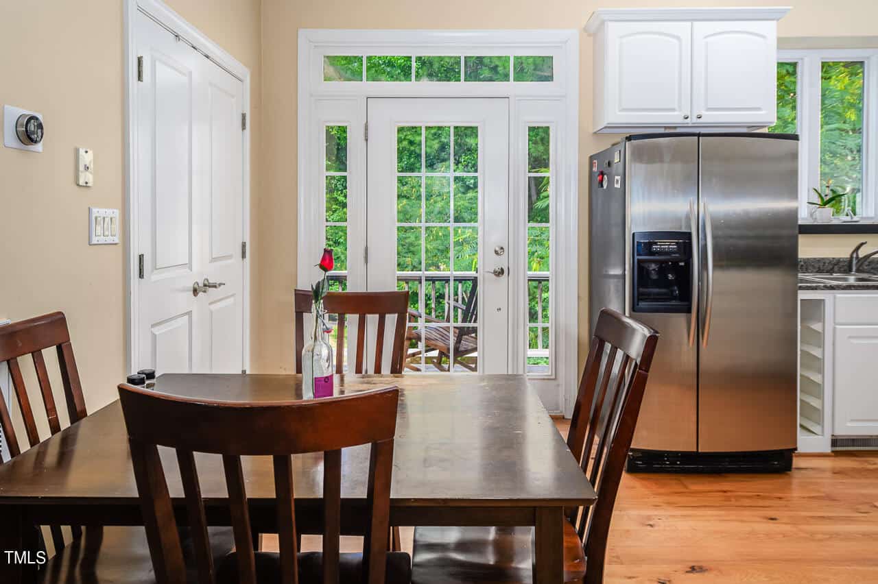 A kitchen with stainless steel appliances, a wooden dining table with four chairs, and a single rose in a vase. Large windows and glass doors in the background offer a view of greenery outside.