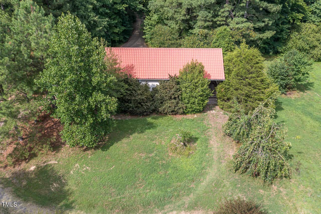 An aerial view of a small house with a red roof surrounded by trees and greenery. A path leads up to the structure through a clearing in the wooded area. The overall scene is lush and verdant.