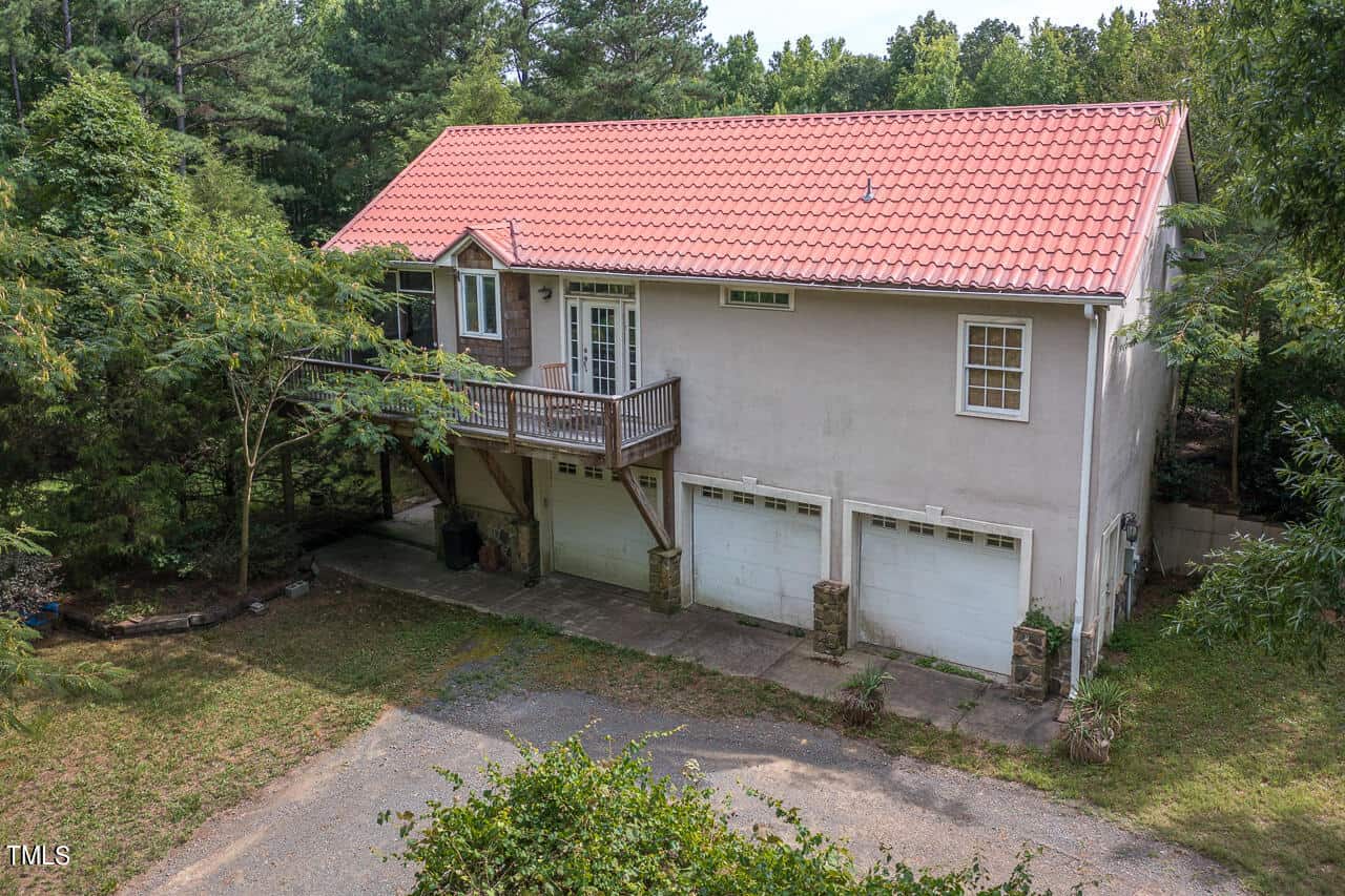 A house with a red tile roof, elevated porch, and three garage doors underneath, surrounded by trees and greenery. The exterior is light-colored with a driveway leading to the garages.
