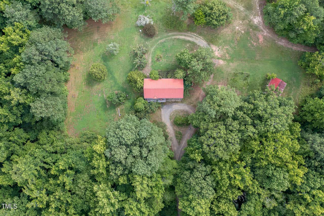Aerial view of a rural landscape at 3158 Tump Wilkins Road in Stem, showcasing a small house with a red roof, encircled by lush green trees. A winding dirt path meanders towards the house, while another small structure peeks from the top right corner.