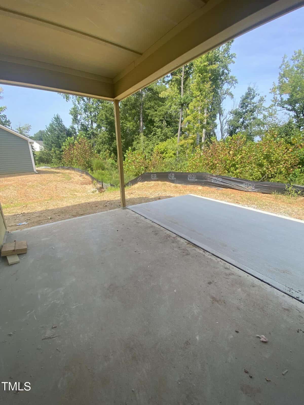 View from a covered patio looking out onto a newly poured concrete slab, with a yard bordered by trees and a black silt fence. The sky is clear and the area appears to be part of a new construction.