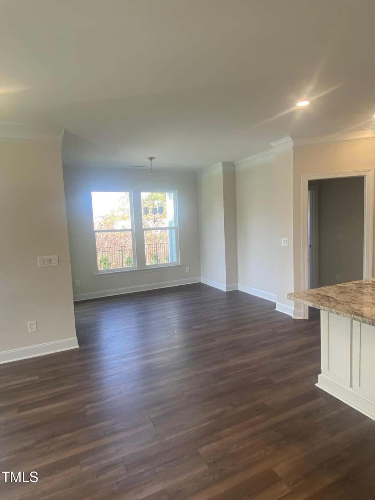 Bright, empty room with large windows, light-colored walls, and dark wood flooring. Part of a kitchen counter with a granite top is visible on the right. Ceiling lights illuminate the space.