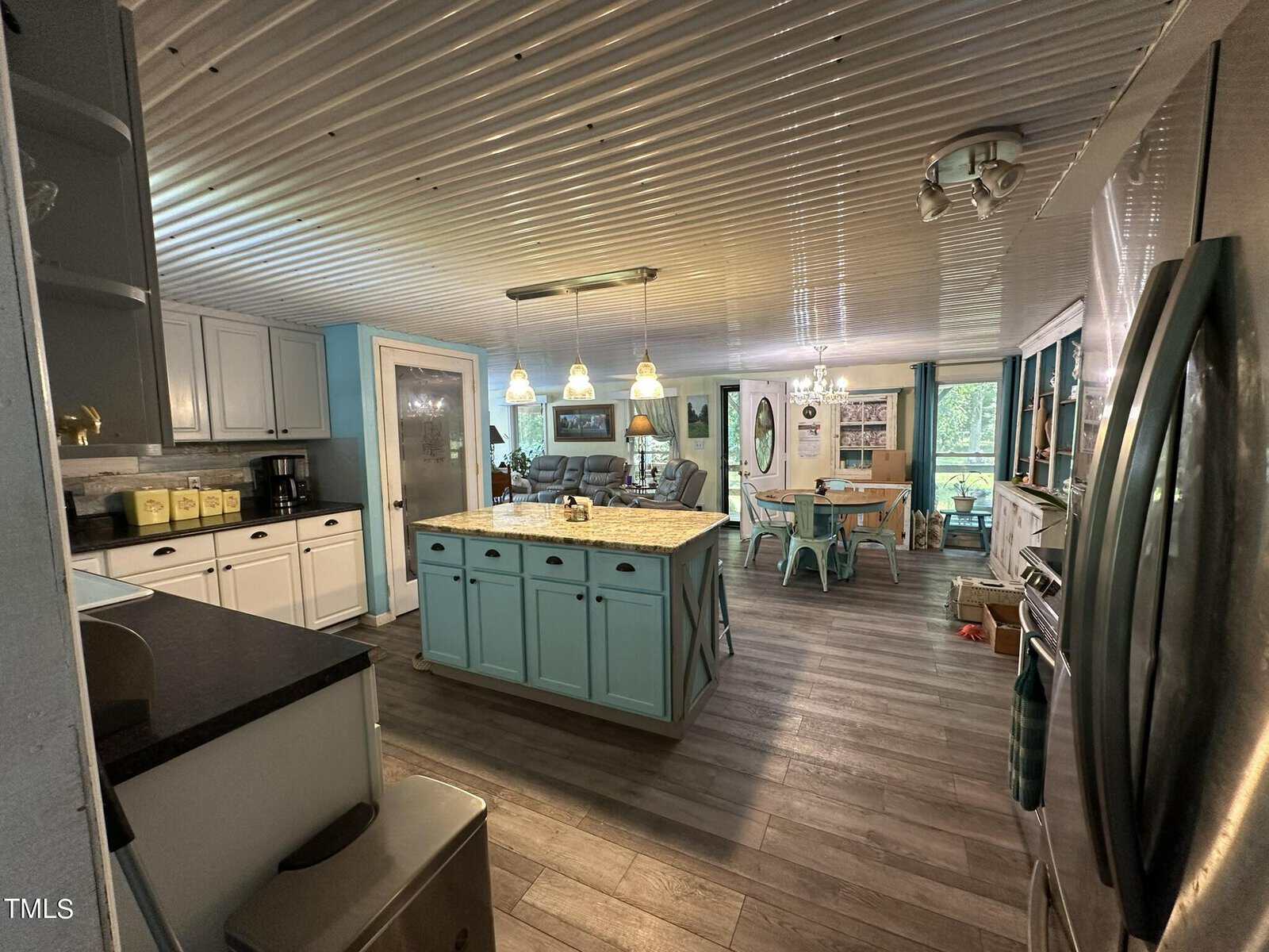 A spacious kitchen with a corrugated ceiling, featuring a central island with blue cabinets. The kitchen is well-lit with pendant lights and has a dining area in the background with a round table and chairs. The floors are wooden.