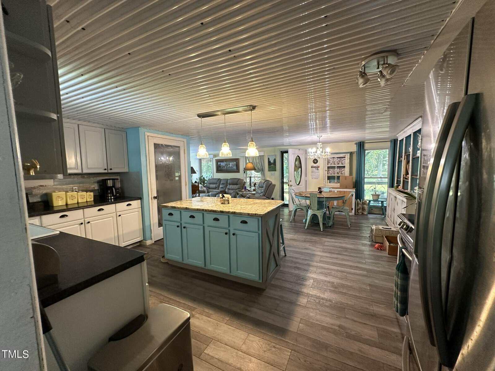 A spacious kitchen with a blue island, modern appliances, and hanging pendant lights. To the right is a dining area with a large window, wooden table, and teal chairs. The room features wood flooring and a corrugated metal ceiling.