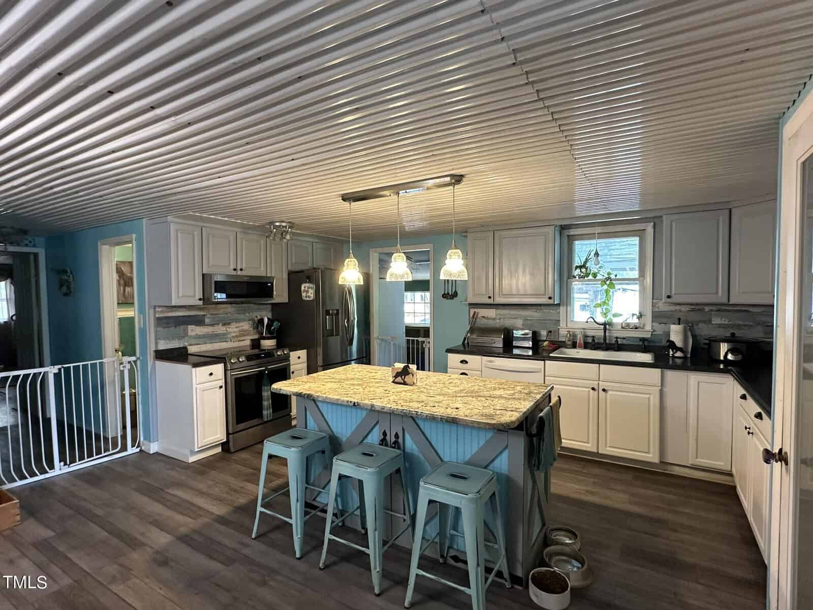 A modern kitchen with a corrugated metal ceiling, white cabinets, and a granite-topped blue island. Three metal stools line the island. Hanging pendant lights illuminate the space, with wood-look flooring and a visible baby gate in the background.