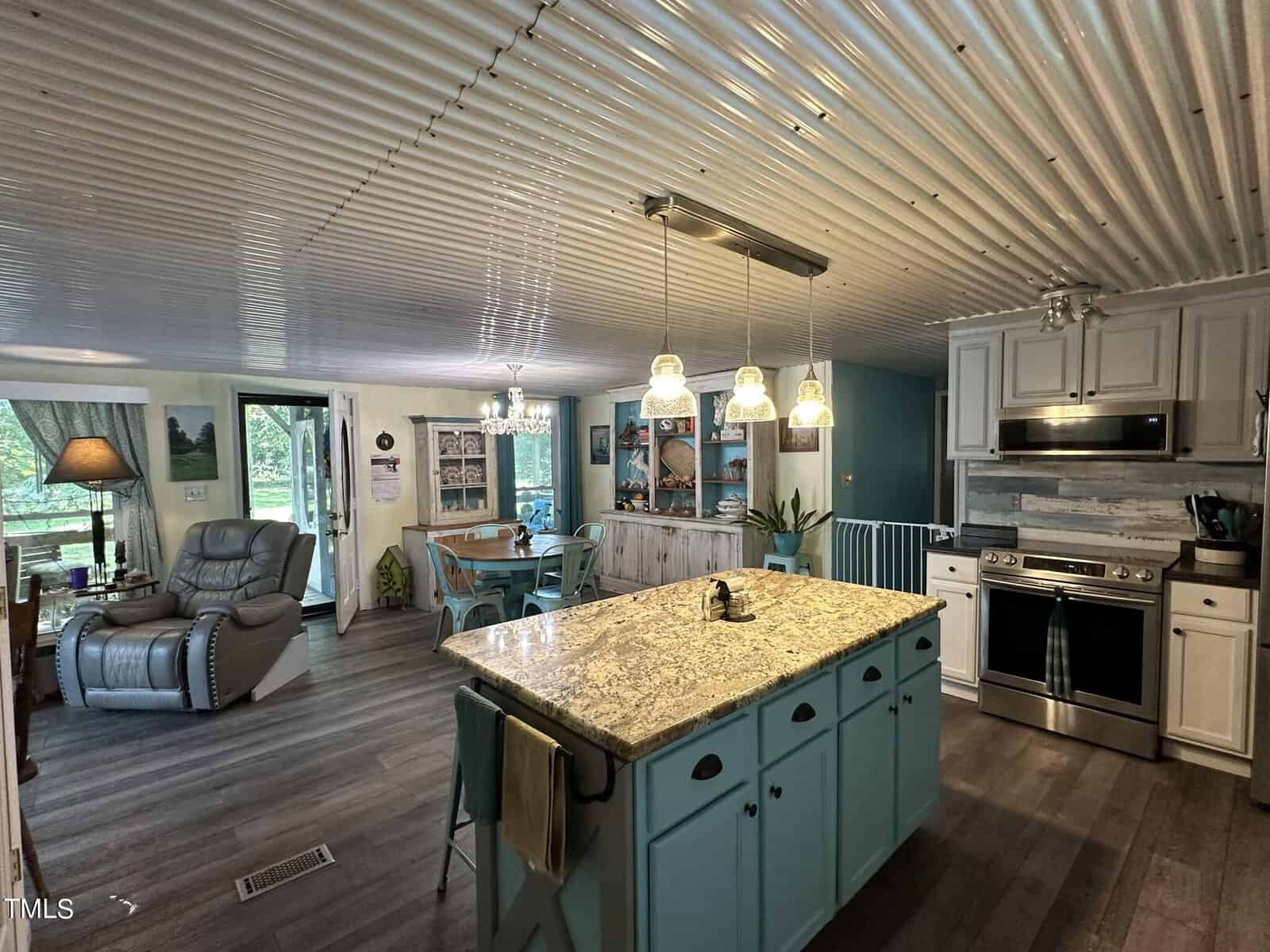 A cozy kitchen with a large granite island, light blue cabinets, and stainless steel appliances. The room features a corrugated metal ceiling, pendant lights, and a view into a living area with armchairs and a window overlooking greenery.
