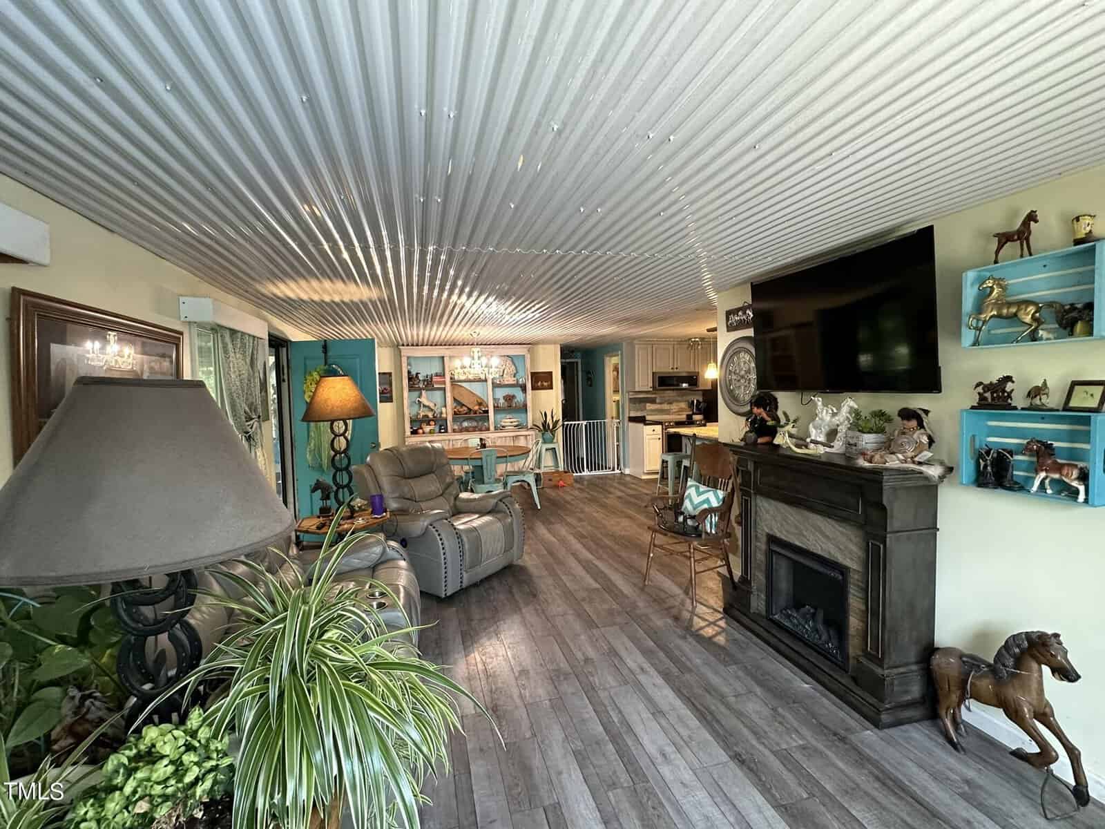 A cozy living room with a corrugated metal ceiling, wooden flooring, and a fireplace. There are decorative horse figurines, a large TV, seating area, and plants. The kitchen is visible in the background.