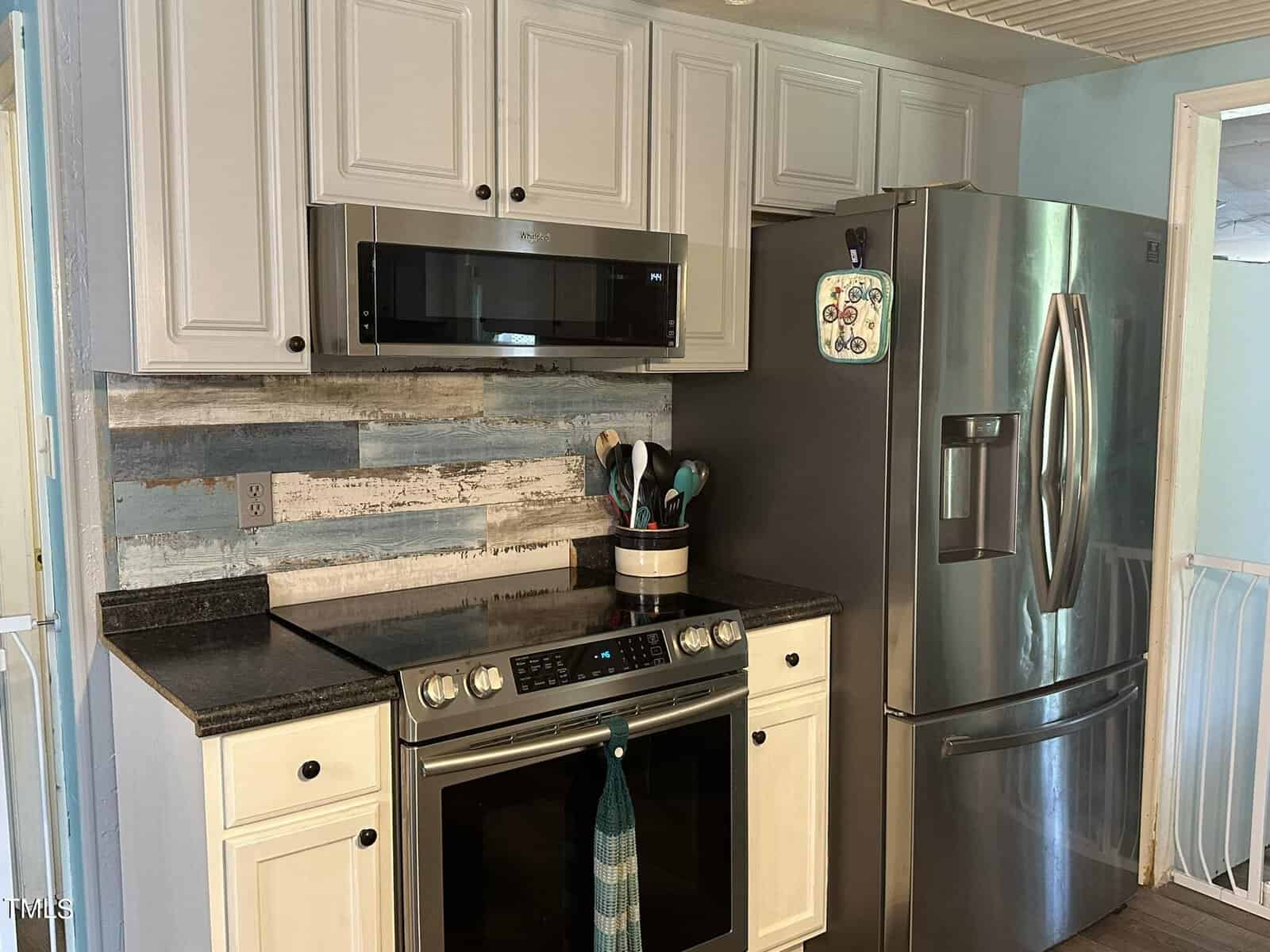 A kitchen featuring a stainless steel refrigerator, stove, and microwave. White cabinets are above the stove, with a rustic wood-patterned backsplash. Utensils are stored in a container beside the stove.