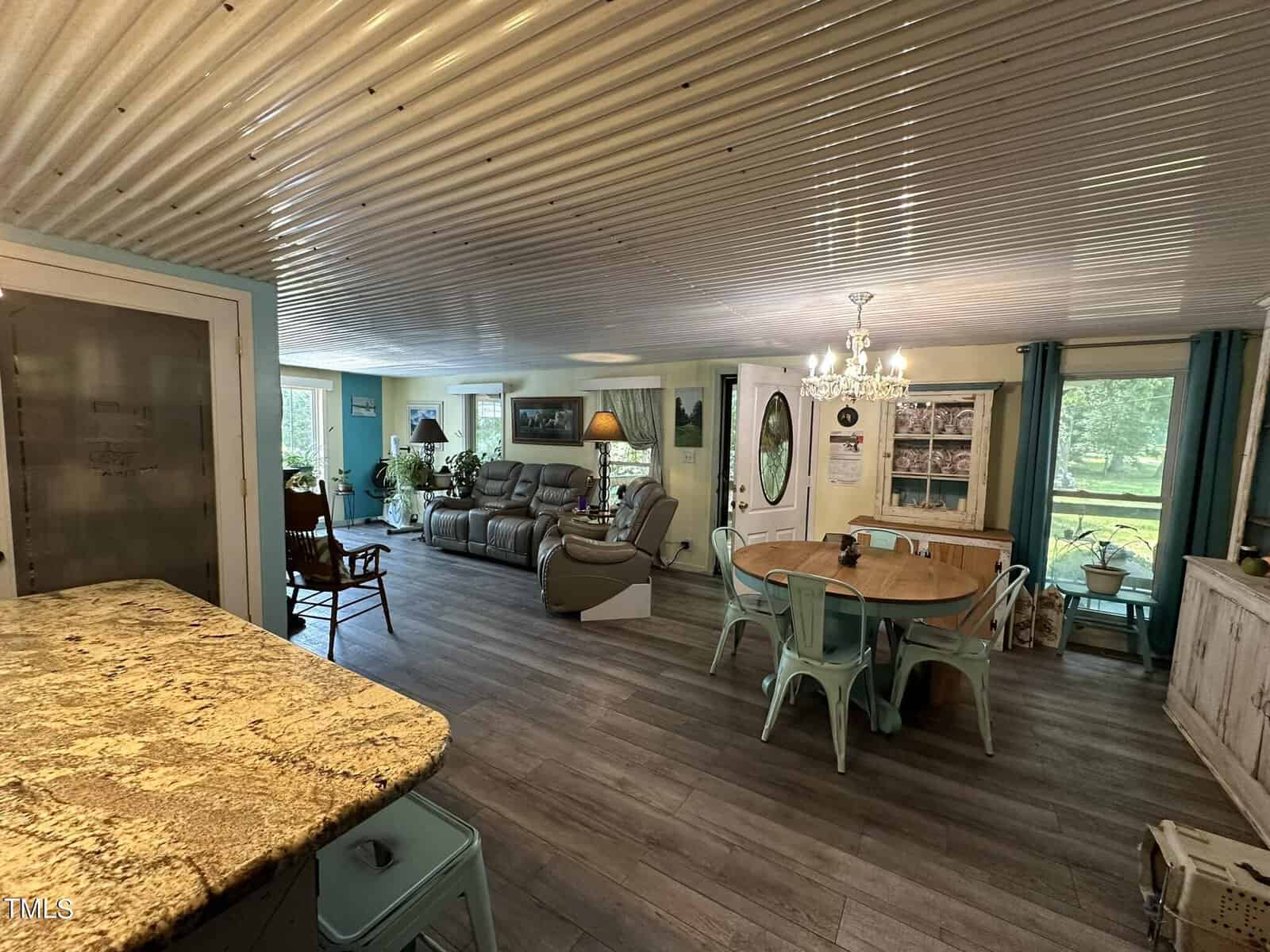 A cozy open-plan living area features a dining table with turquoise chairs, a granite kitchen countertop, and a comfortable seating area with a couch. Natural light streams in from several windows, and a chandelier hangs above the dining space.