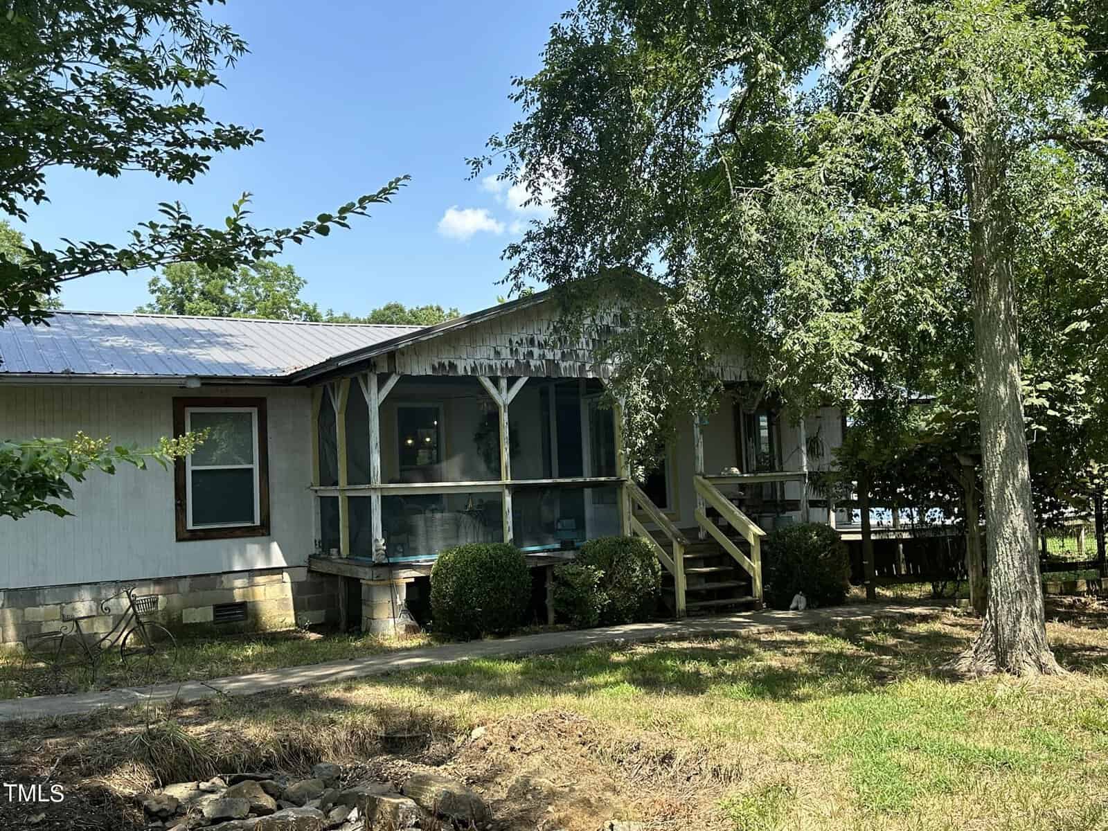 A single-story house with a tin roof and a wooden porch. There are several trees around the yard, providing shade. Steps lead up to the porch, which is partially enclosed. The house is surrounded by grass and a small garden area.