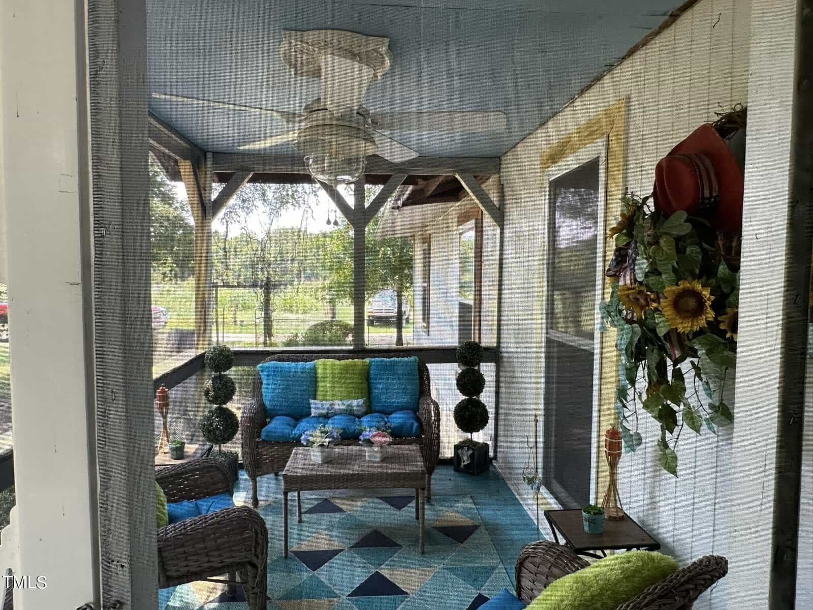 Cozy screened porch with wicker furniture, blue and green cushions, patterned floor rug, ceiling fan, and decorative plants. A wall features a wreath with sunflowers and hats. Scenic outdoor view in the background.