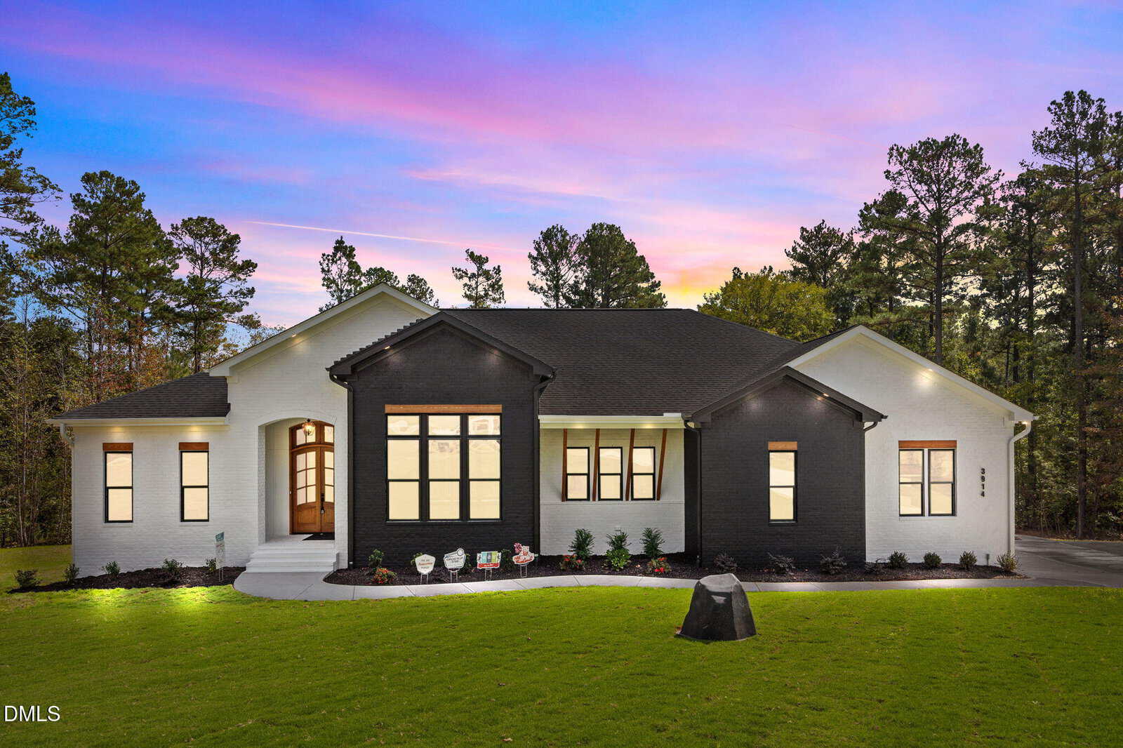 A modern single-story house at 3914 Sage Court in Wake Forest features a sleek black and white exterior, set on a green lawn among trees. Large windows glow from within as the sky turns pink and purple at sunset.