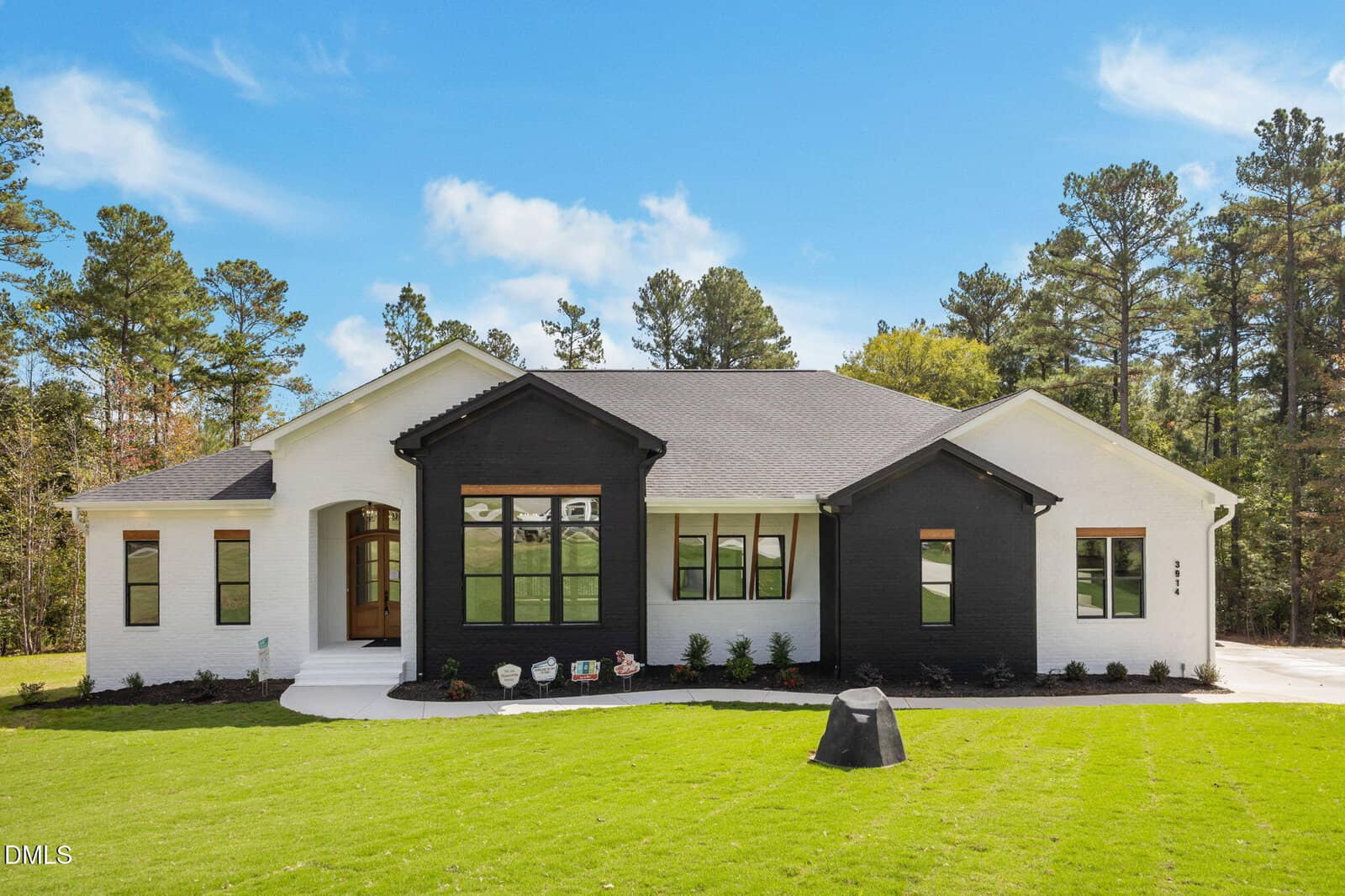 A modern single-story house with white and black exterior paint, large windows, and a well-maintained green lawn. The house is surrounded by trees under a blue sky with some clouds.