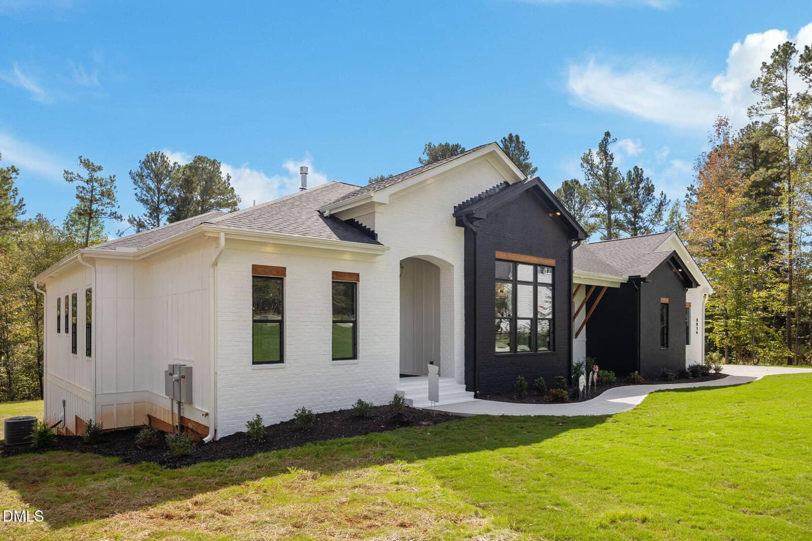 A modern single-story house with white brick exterior and a black accent section. The home has multiple windows, a sloped roof, and a landscaped yard with green grass and surrounding trees under a blue sky.