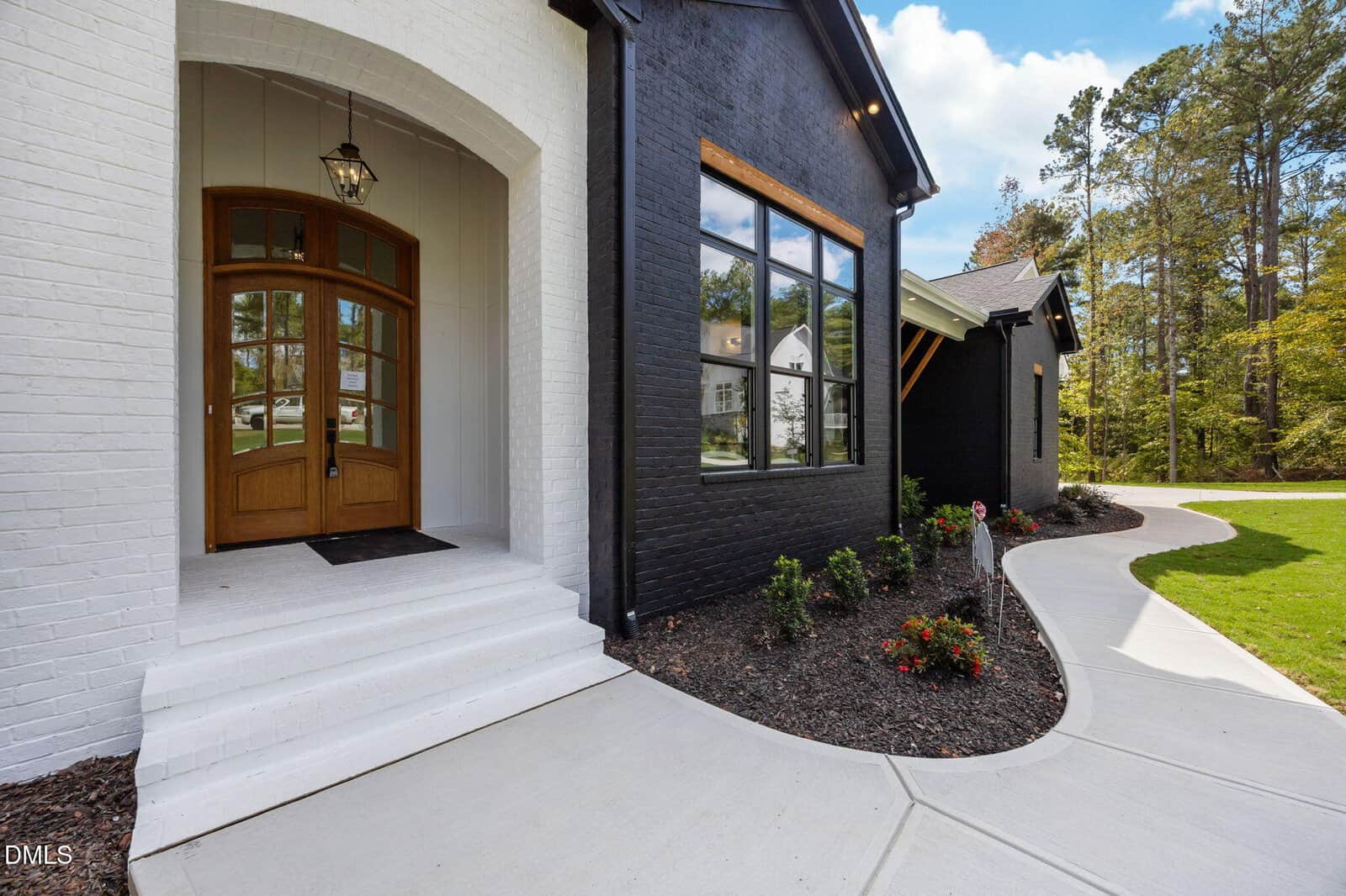 A modern house exterior features a white brick entrance with wooden double doors, adjacent to a black brick facade with large windows, landscaped shrubs, and a curved concrete walkway.