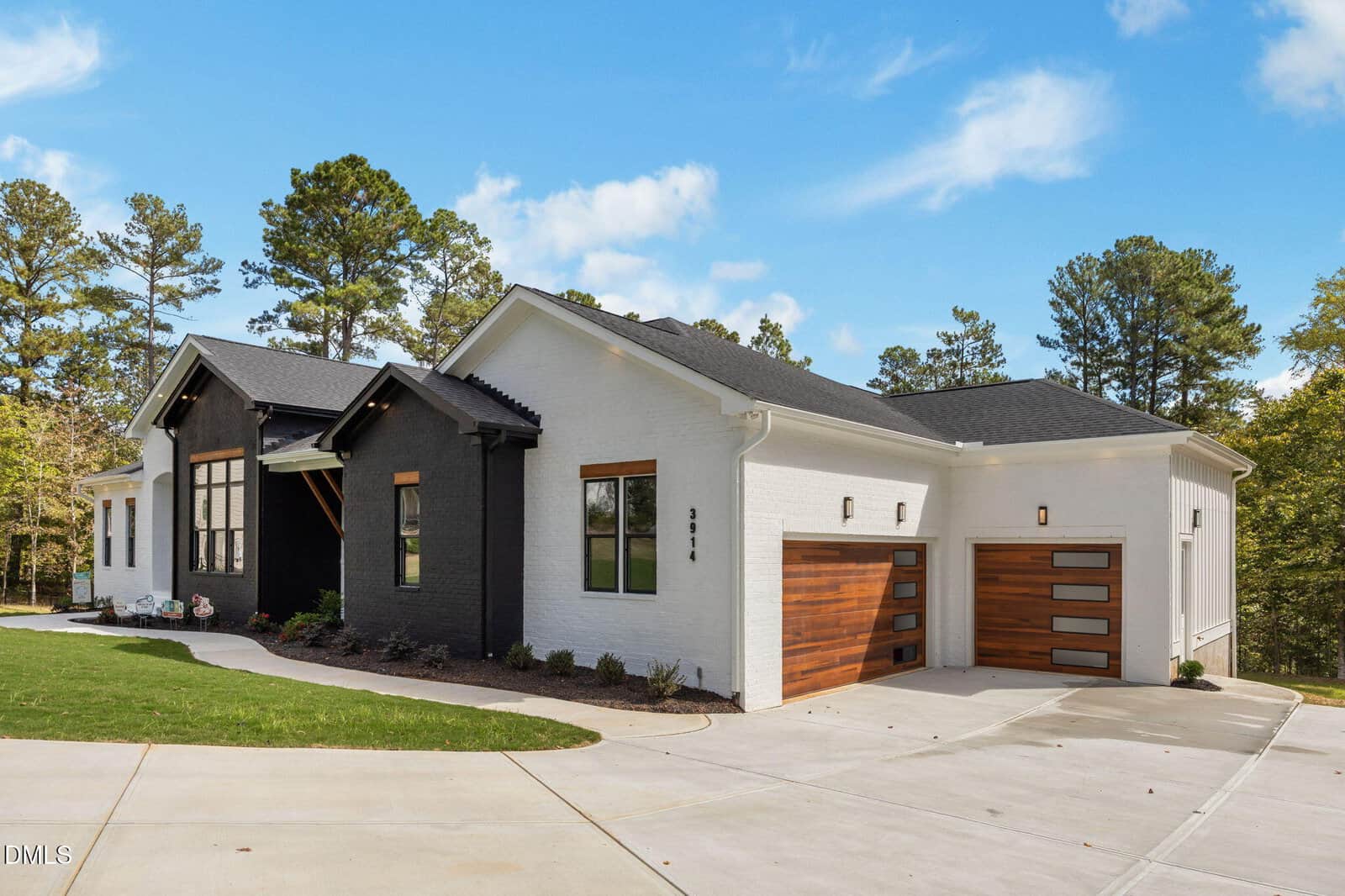 A modern single-story house with white and black exterior walls, large front windows, and two wooden garage doors, set against a backdrop of tall trees and a clear blue sky.