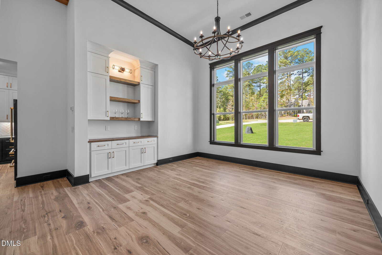 A bright, empty room with large windows, light wood flooring, white walls, built-in cabinets with shelves, and a black chandelier hanging from the ceiling. Green lawn and trees are visible outside.