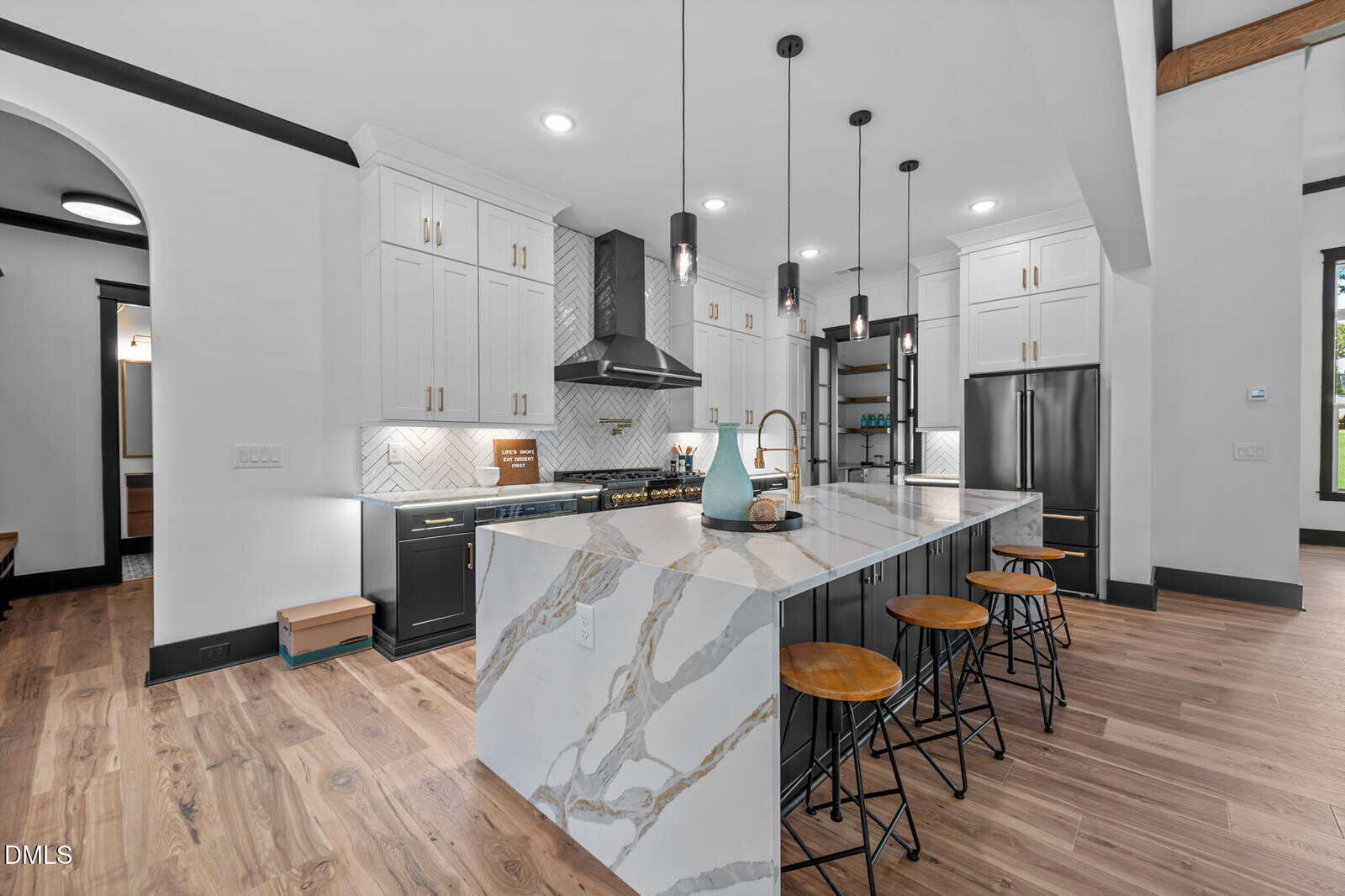 Modern kitchen with white cabinets, black lower cabinets, stainless steel appliances, a marble waterfall island with three wooden stools, pendant lighting, and wood floors. Decorative vases sit on the island.
