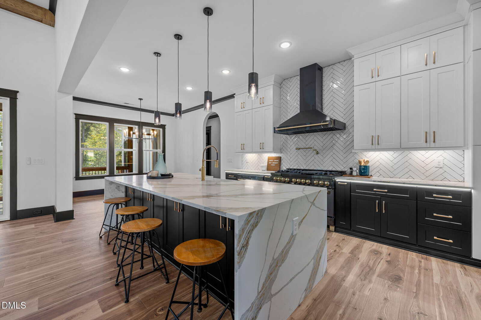 Modern kitchen with a large marble island, black and white cabinetry, bar stools, stainless steel appliances, and a herringbone tile backsplash. Pendant lights hang above the island, and wood flooring is visible.