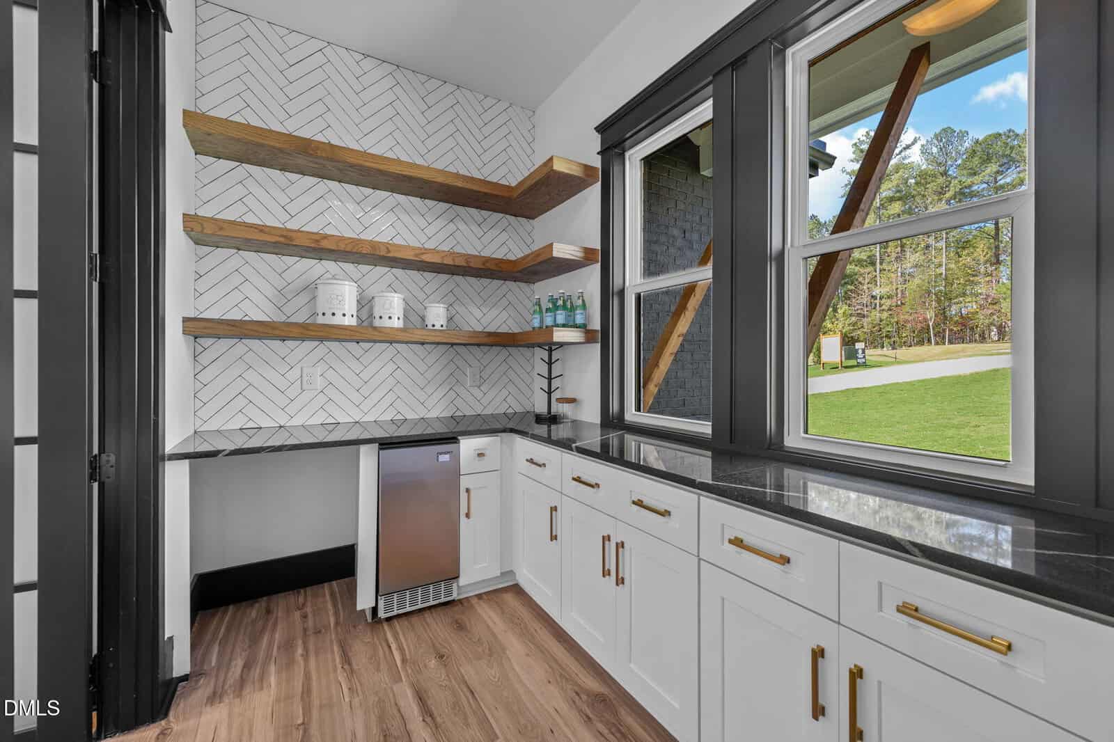 A modern kitchen with white cabinets, black countertops, wood shelves, herringbone tile backsplash, a mini fridge, canisters, bottled drinks, and large windows overlooking a green outdoor area.