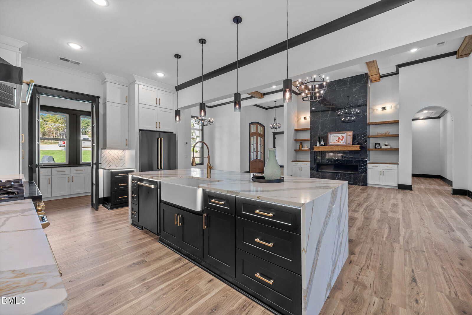 Modern kitchen with black and white cabinetry, marble countertops, a farmhouse sink, pendant lighting, stainless steel appliances, light wood flooring, and an open layout with a fireplace and built-in shelves in the background.