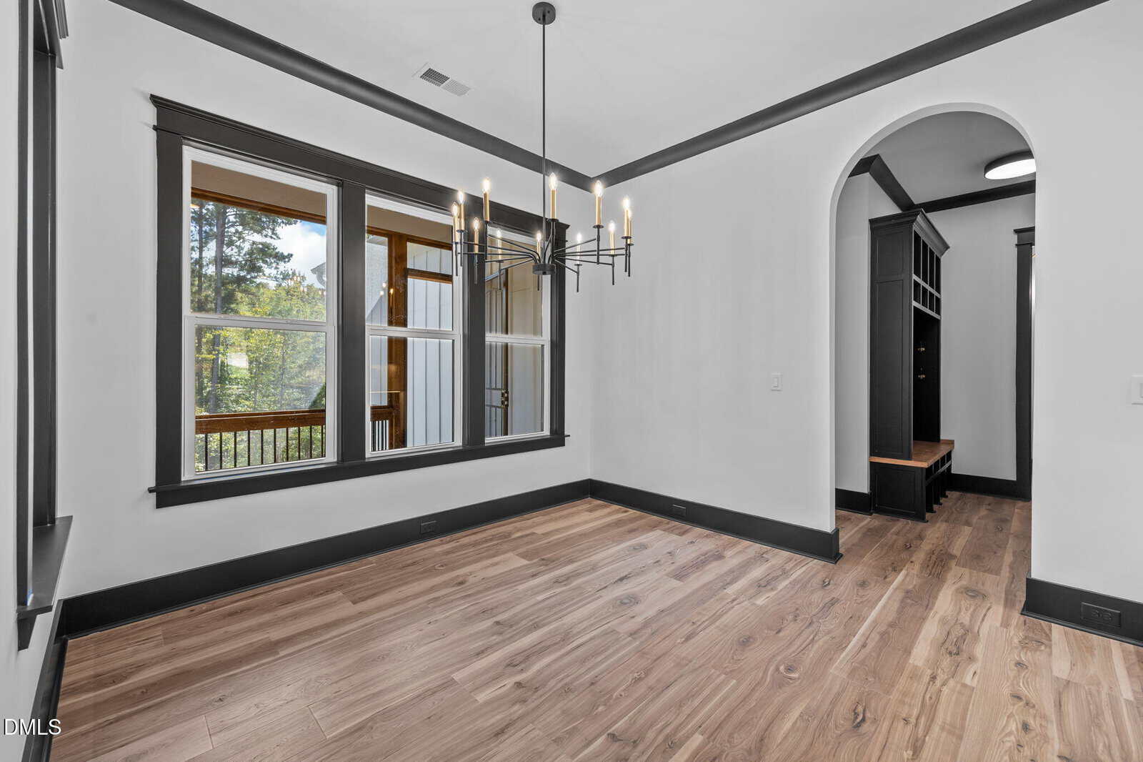 A bright, empty room with light wood floors, white walls, black trim, a modern black chandelier, large windows, and an arched doorway leading to another area with built-in shelving and a bench.