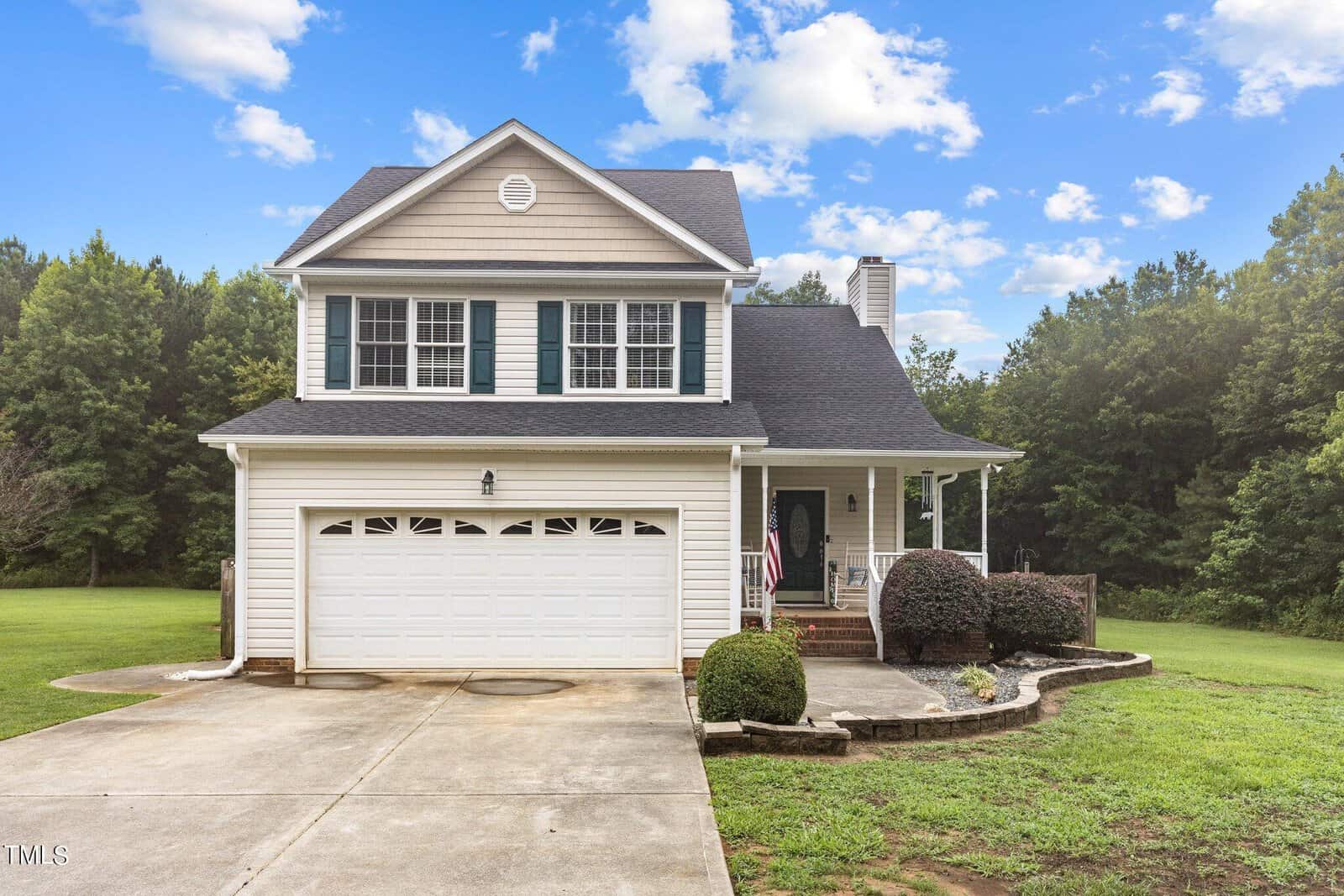 Nestled on 3690 Mabel Lane, this charming two-story suburban house boasts beige siding and black shutters. It features a welcoming front porch and a two-car garage, all surrounded by a well-maintained lawn with trees swaying gently under the partly cloudy sky.