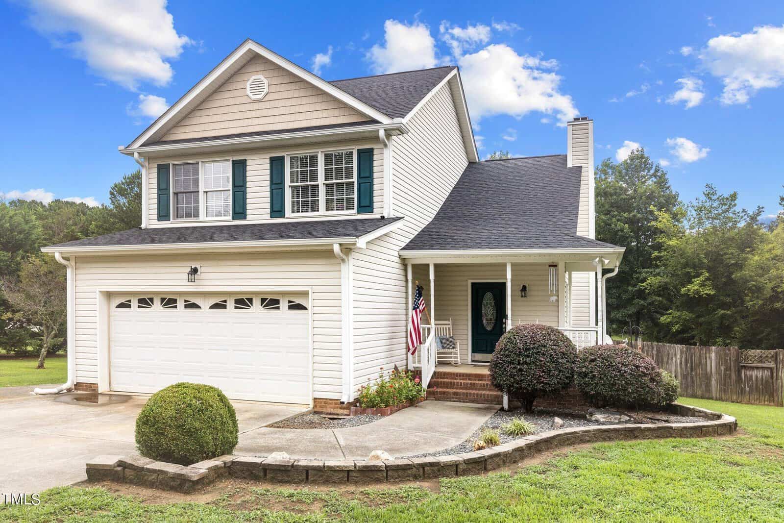 A two-story suburban house with beige siding and a dark shingled roof. It features a double garage, green shutters, and a front porch with an American flag. The yard is landscaped with shrubs and a stone-bordered walkway.