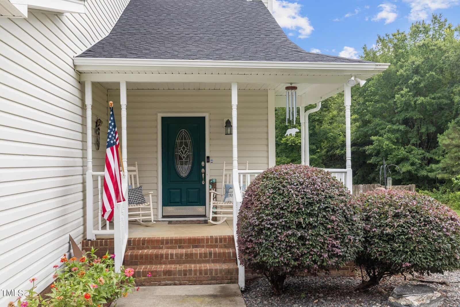 A cozy porch with a green door, rocking chair, and wind chime. An American flag hangs on a post near neatly trimmed bushes. The house has white siding and brick steps. The background features lush greenery and a partly cloudy sky.