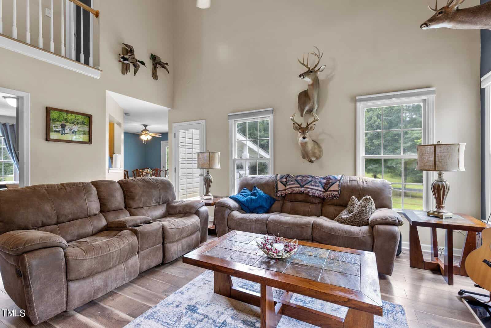 A cozy living room with brown sofas, a wooden coffee table, and a patterned rug. The walls have mounted deer heads as decor. Large windows let in natural light, and a door opens to a lush green view outside.