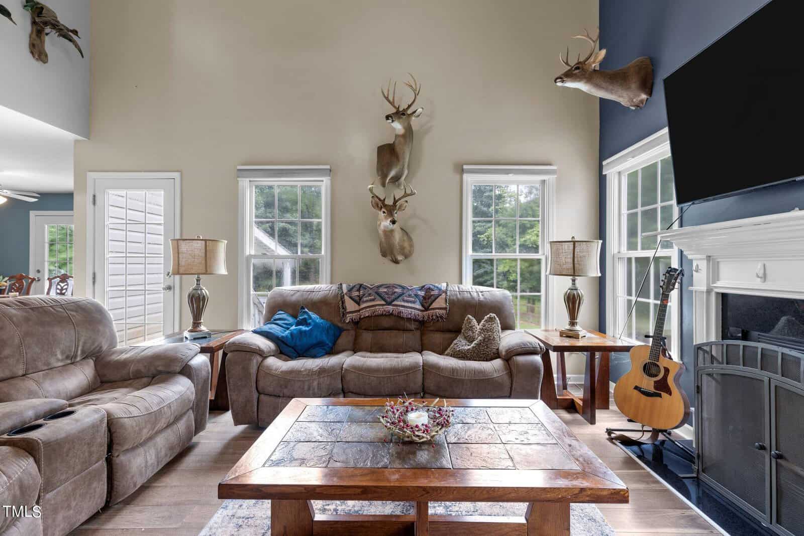 A cozy living room with tall ceilings, featuring a brown leather couch and recliner. Three mounted deer heads adorn the wall. A fireplace with a TV above is on the right, and a guitar rests beside it. Large windows offer a view of greenery outside.
