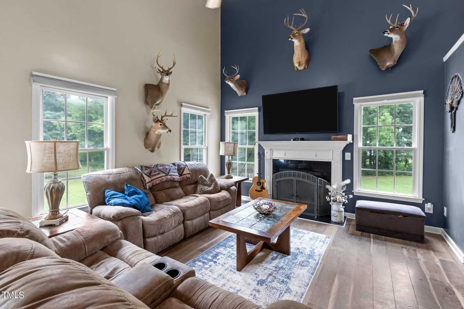 A cozy living room with two sofas, a wooden coffee table, and a large TV above a fireplace. The walls feature mounted deer heads. Two windows provide natural light, and a guitar rests by the fireplace. A blue and white rug covers the wooden floor.