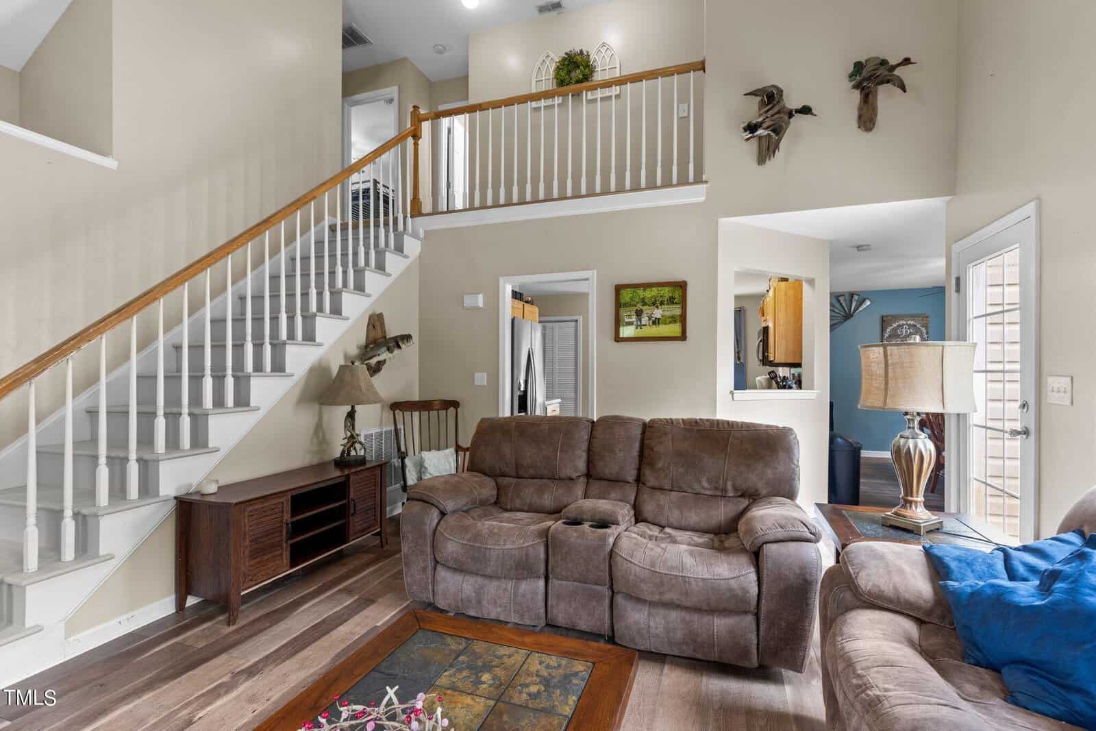 A cozy living room with a brown reclining sofa and loveseat on wood flooring. A staircase with a white railing leads to an upper floor. The room is decorated with wall art and has a lamp on a wooden console table beneath the stairs.