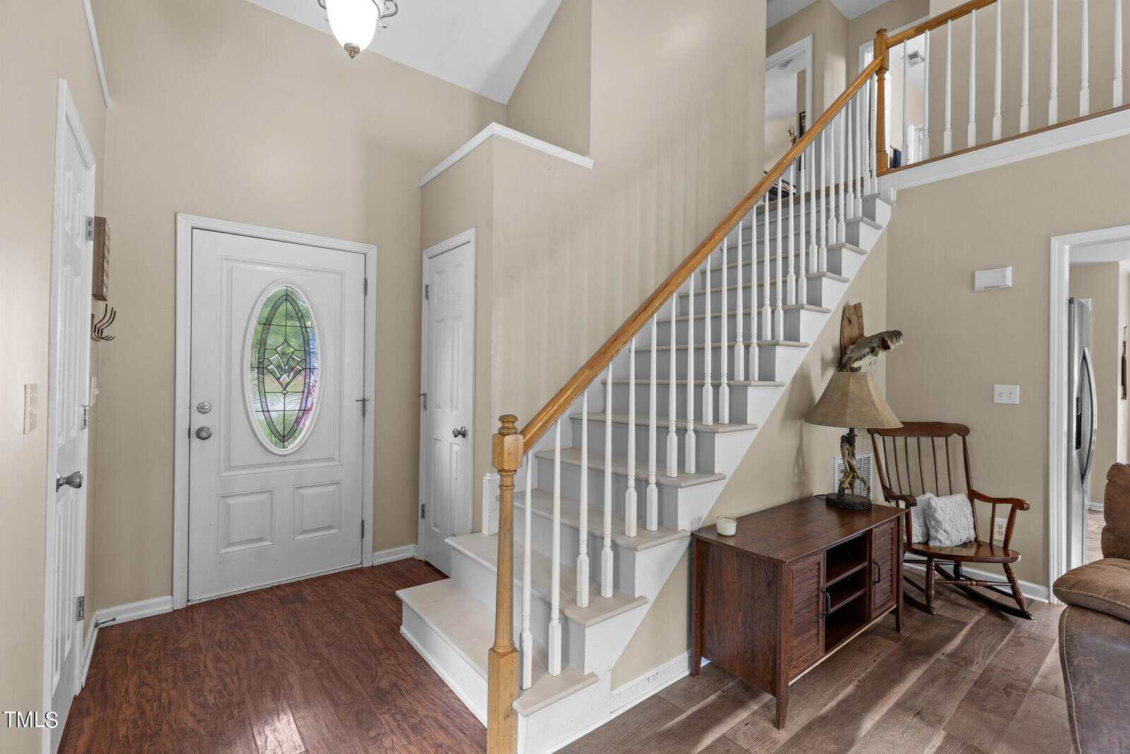 A home entryway with a staircase on the right, featuring white steps and a wooden handrail. A door with an oval stained glass window is on the left. Hardwood floors and light-colored walls complete the space. A wooden chair and side table are near the stairs.