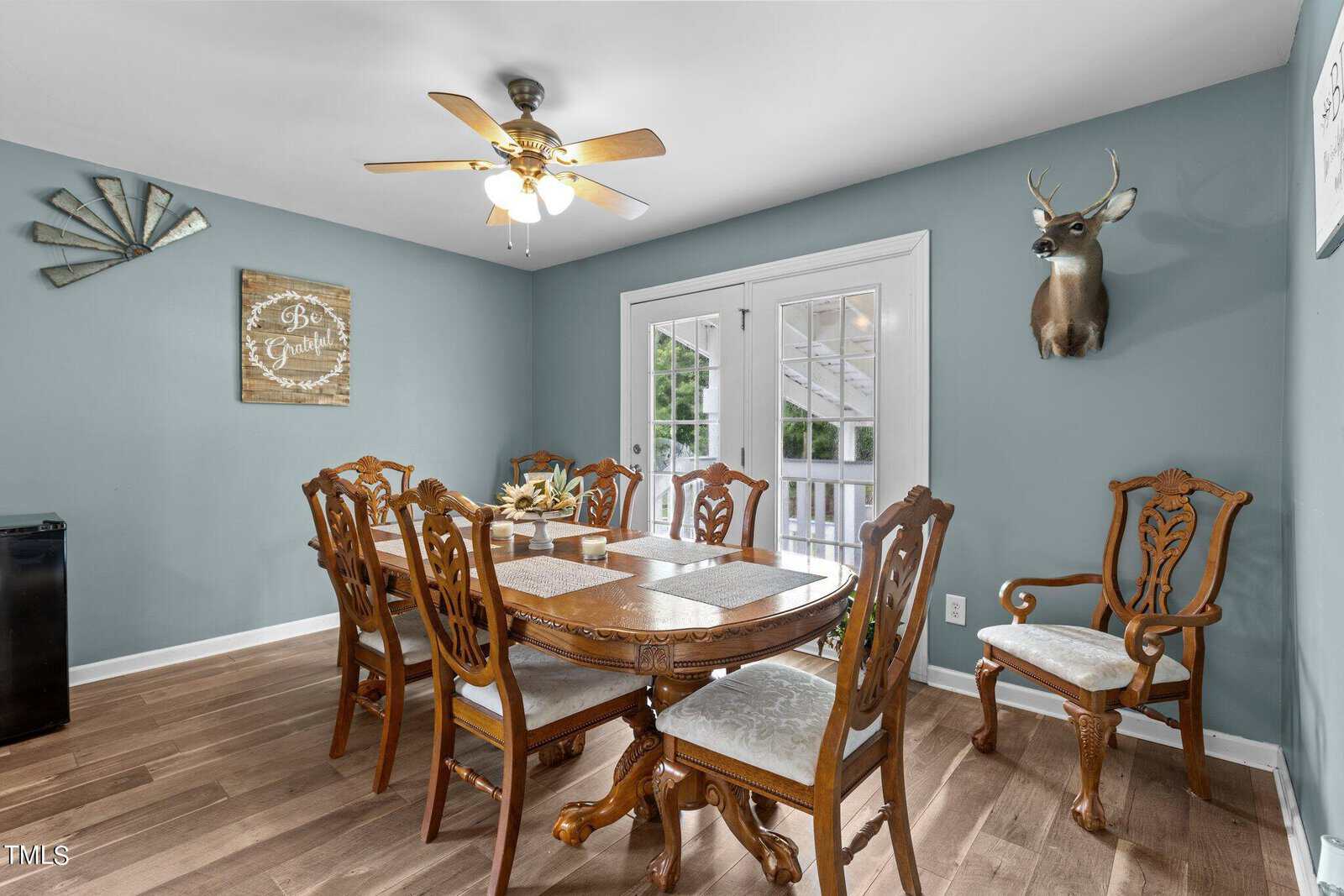 Dining room with a large wooden table and eight chairs. A ceiling fan hangs above. French doors lead outside, and a mounted deer head is on the wall. The walls are painted teal, and there's a rustic sign saying "Be Grateful.