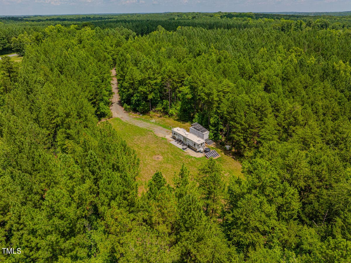 Aerial view of a small cabin-like structure with solar panels on the roof, nestled in a clearing surrounded by dense forest. A narrow dirt path leads to the building. Lush greenery and a clear blue sky are visible in the background.