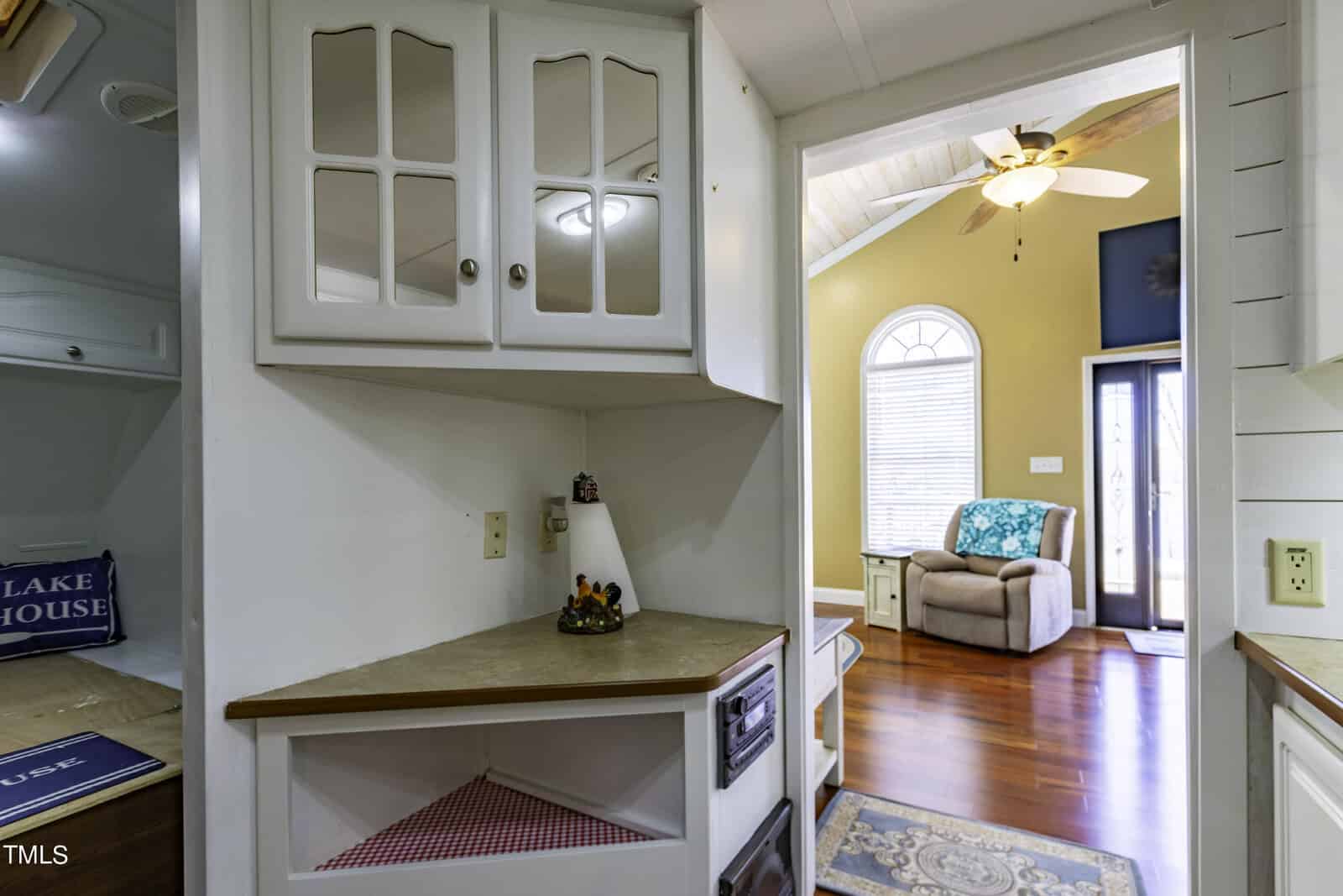 A cozy living area with warm wood floors, a ceiling fan, and arched windows is visible through a doorway; the foreground shows a small corner kitchen with white cabinets and beige countertops.