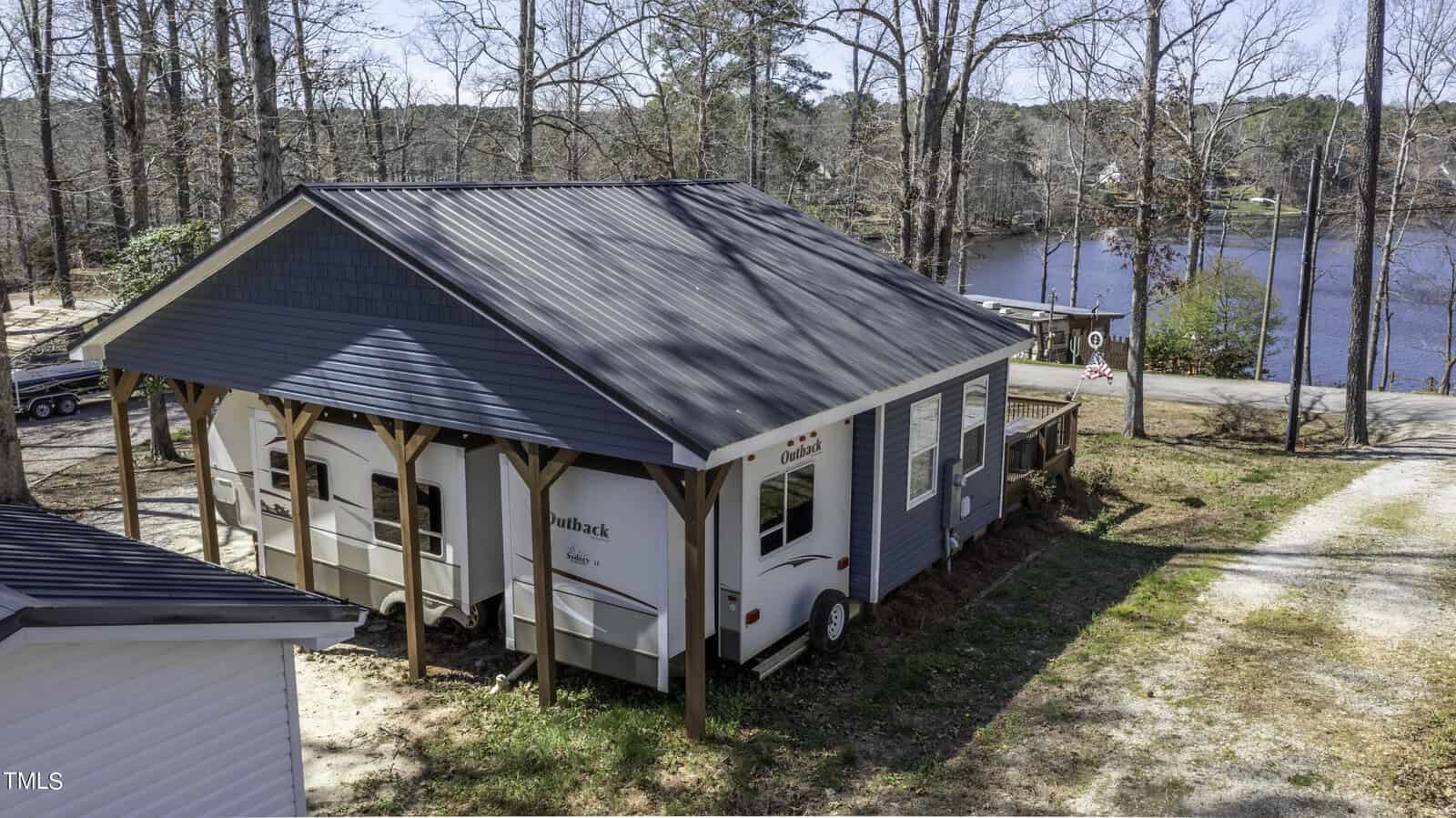 A small blue house with a metal roof built around a travel trailer sits near a lake, surrounded by tall trees and a dirt driveway leading to the water's edge.
