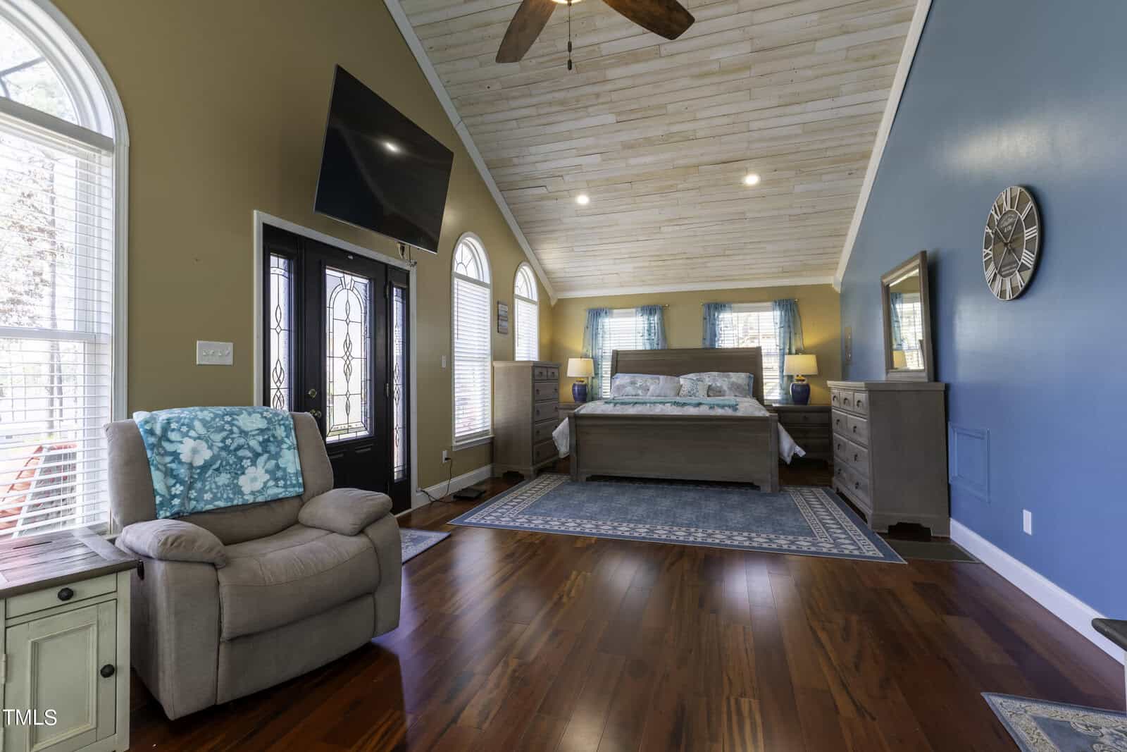 Spacious bedroom with wood floors, a large bed, blue accent wall, vaulted ceiling with fan, armchair, dressers, TV, and double doors with windows. Blue curtains and rugs add color to the room.