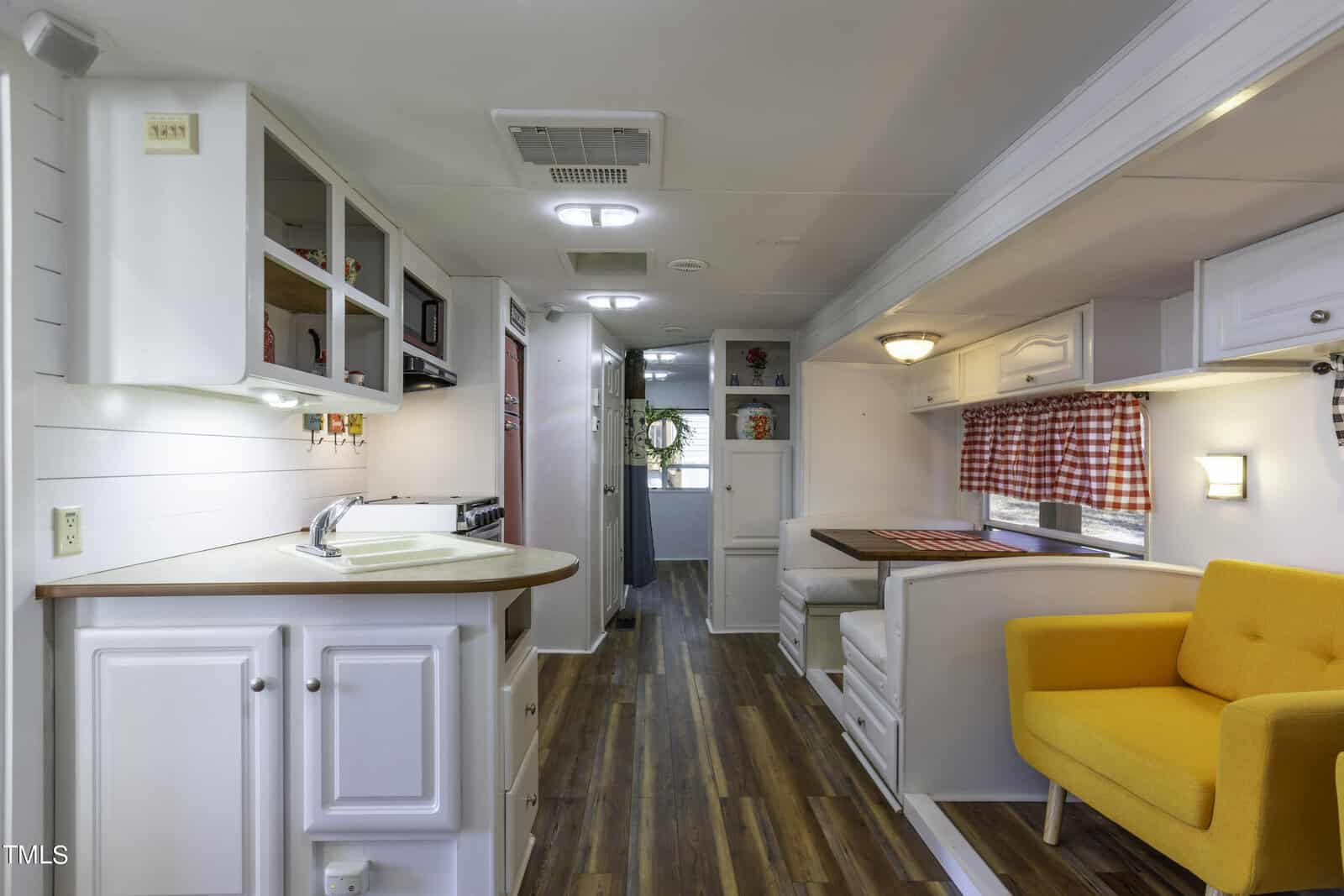 Bright, modern RV interior with white cabinets, a wood floor, a built-in dining area with red checkered curtains, and a vibrant yellow armchair. Natural light streams in from windows at the far end.