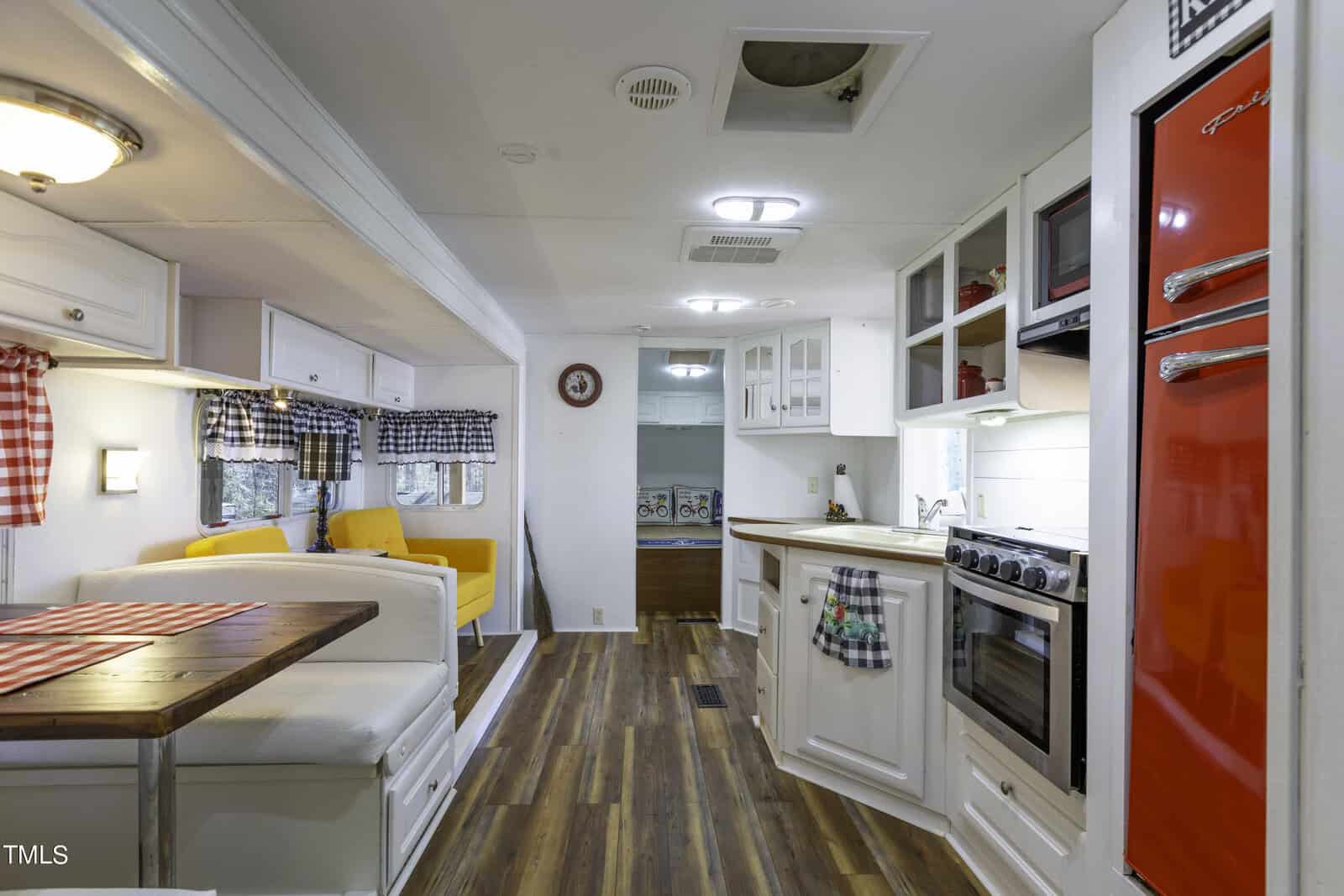 Interior of a retro-style camper with a dining booth, wood flooring, yellow sofa, white cabinetry, checkered curtains, and a red refrigerator. The kitchen area includes a stove, oven, and sink.