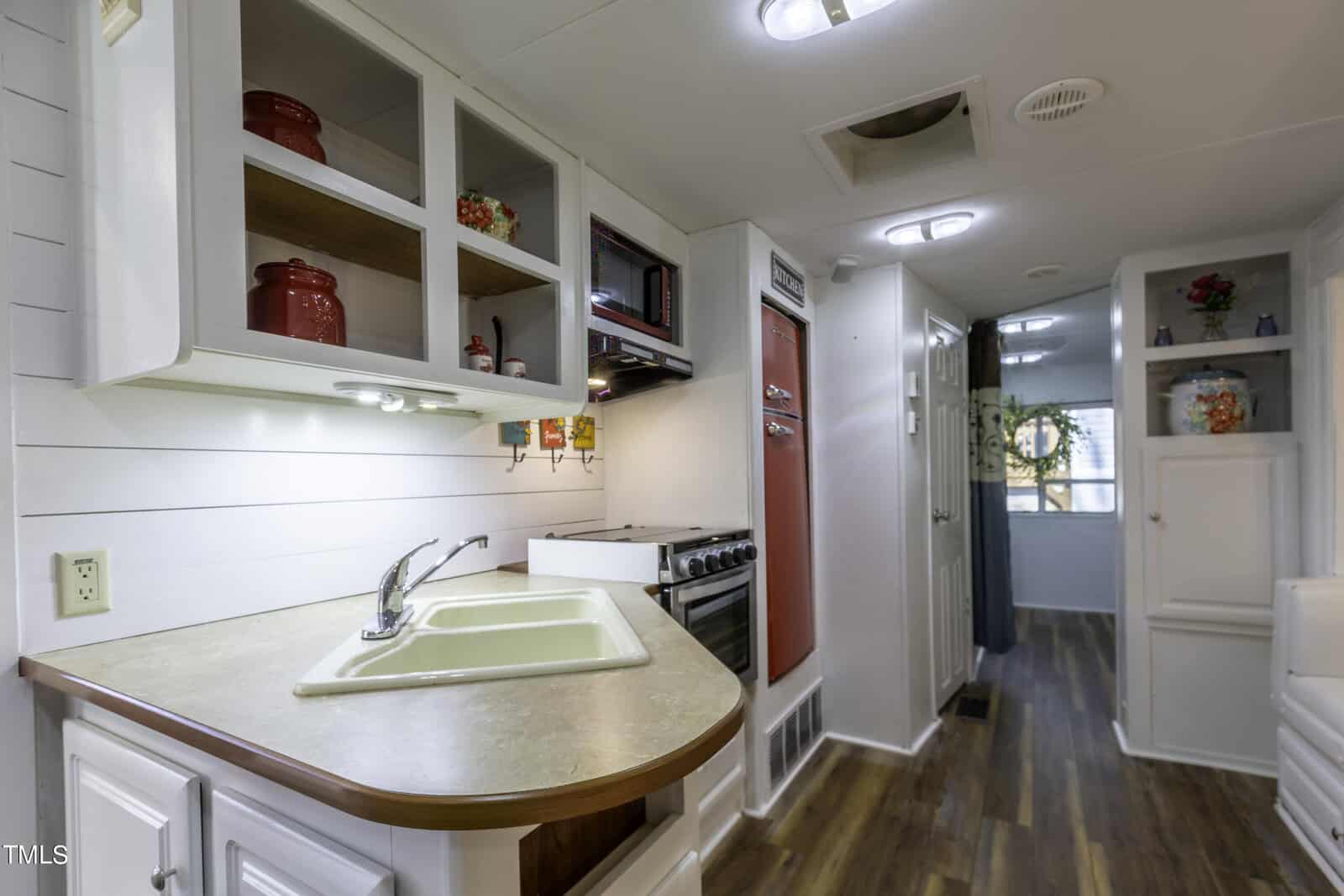 A small kitchen with white cabinets, wood countertops, a white sink, red appliances, and shelves holding red jars. The space has wood flooring and leads to a hallway with a circular window at the end.